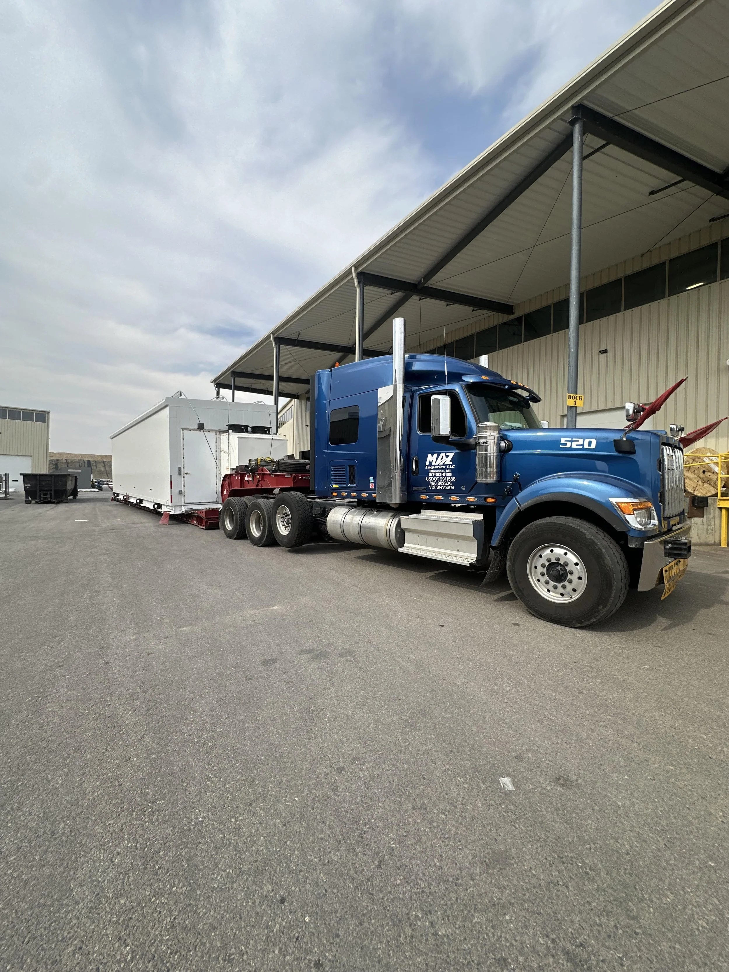 A blue semi-truck parked outside a warehouse, attached to a large white trailer, with a partly cloudy sky overhead.