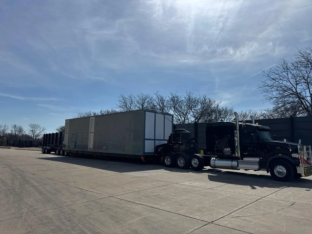 A black semi-truck with a large trailer parked in an open lot with trees and a clear sky in the background.