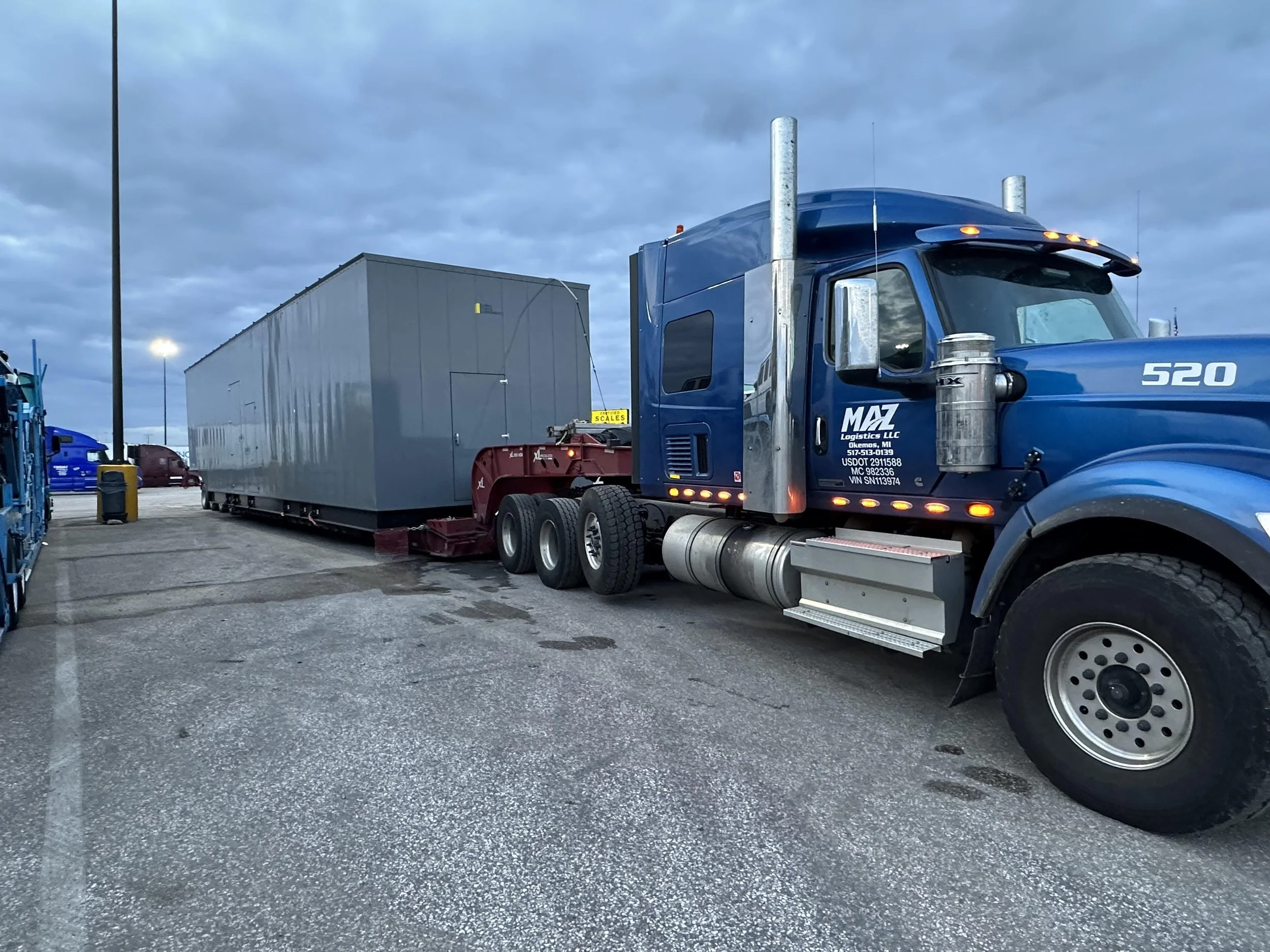 A large blue semi-truck hauling a grey container in a parking lot during dusk or early evening.