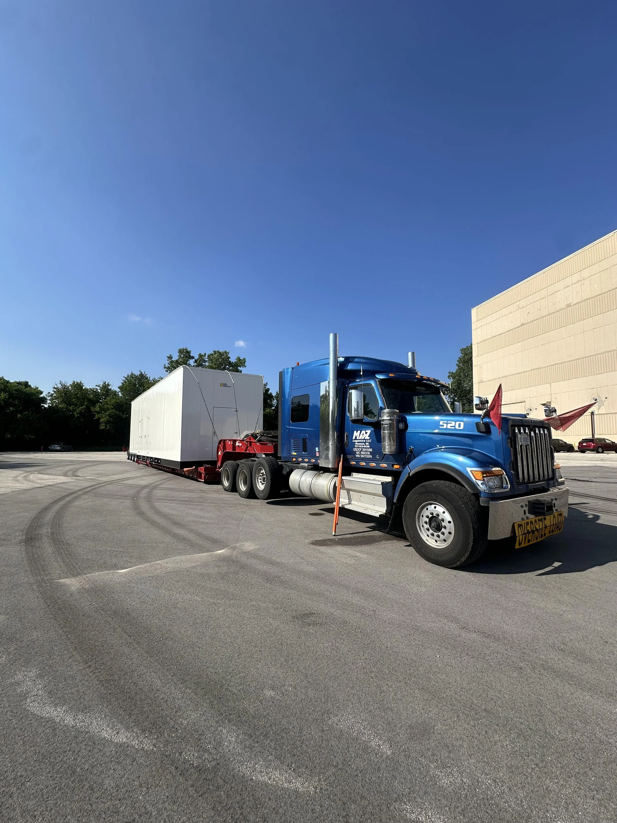 A blue semi-truck with a cargo trailer attached, parked on a paved lot near a large beige building, with trees in the background and a clear blue sky overhead.