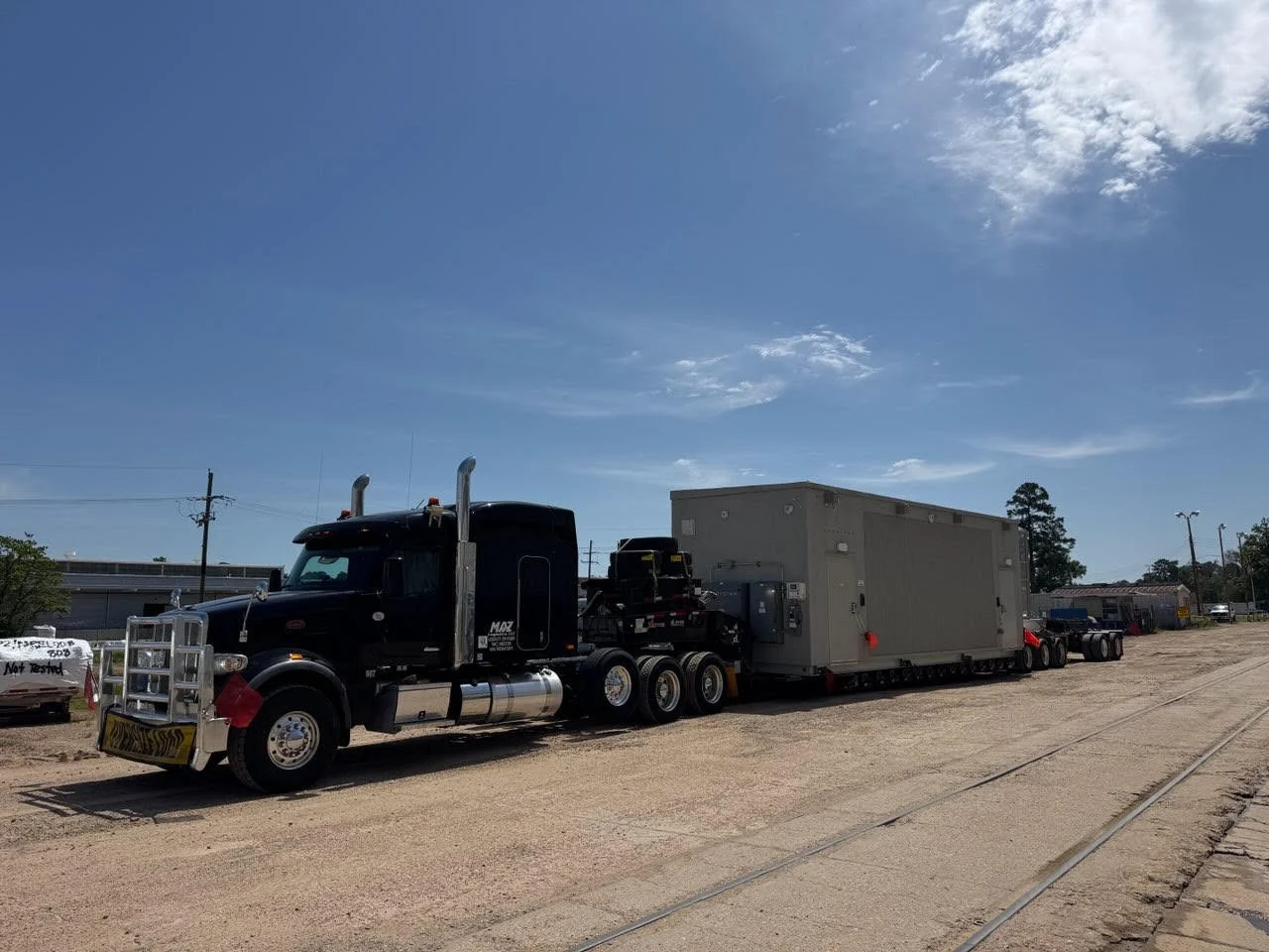 A large black semi-truck with a gray trailer parked on a dirt lot under a partly cloudy sky. The truck is equipped with a grille guard and exhaust stacks, and there are utility boxes and equipment mounted on the trailer.