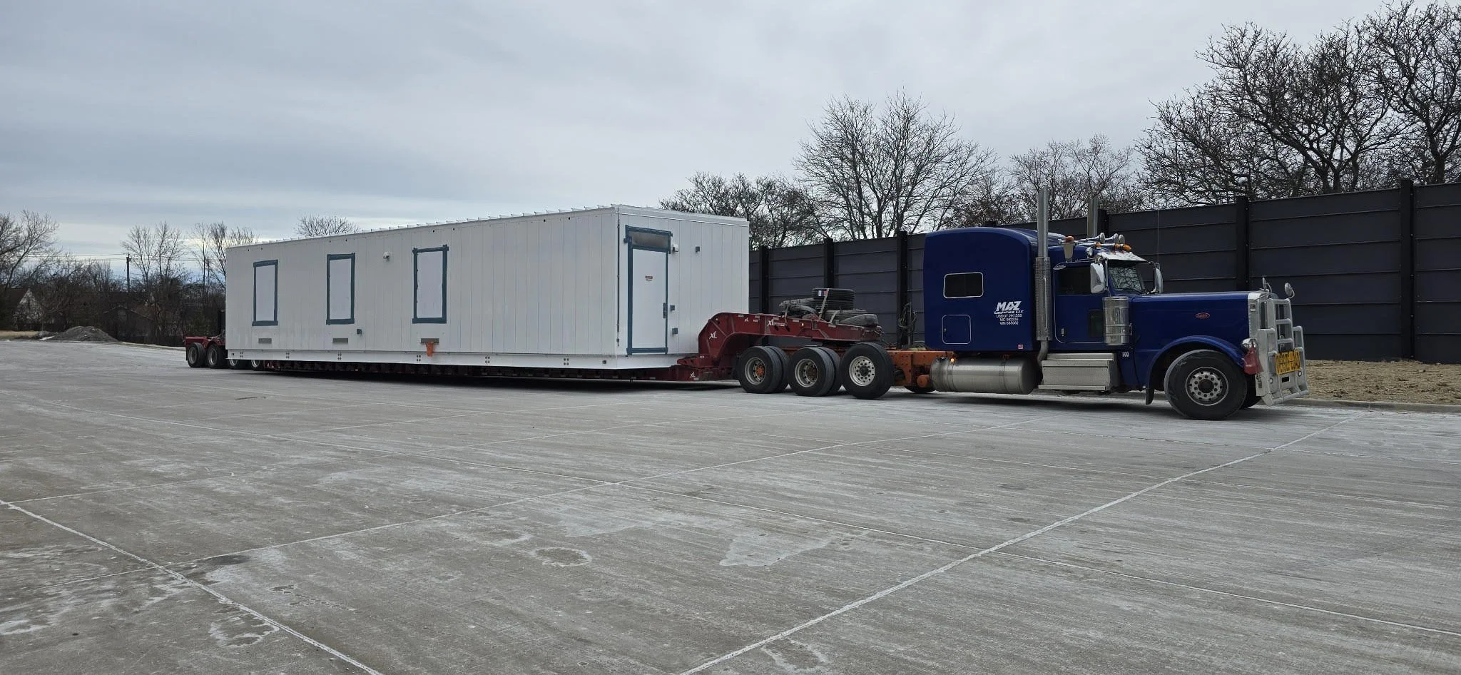 A blue semi-truck hauling a white, prefabricated building or container on a flatbed trailer on a paved lot with a black fence and leafless trees in the background.