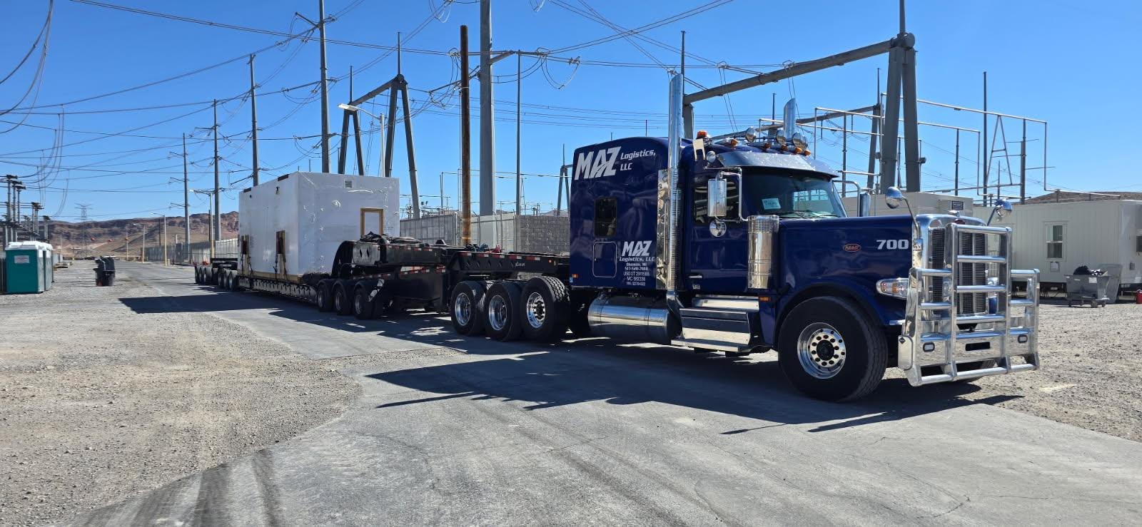 A large Kenworth semi-truck with a blue cab and chrome grille is parked on a gravel lot under power lines, hauling a flatbed trailer with an oversized white structure.