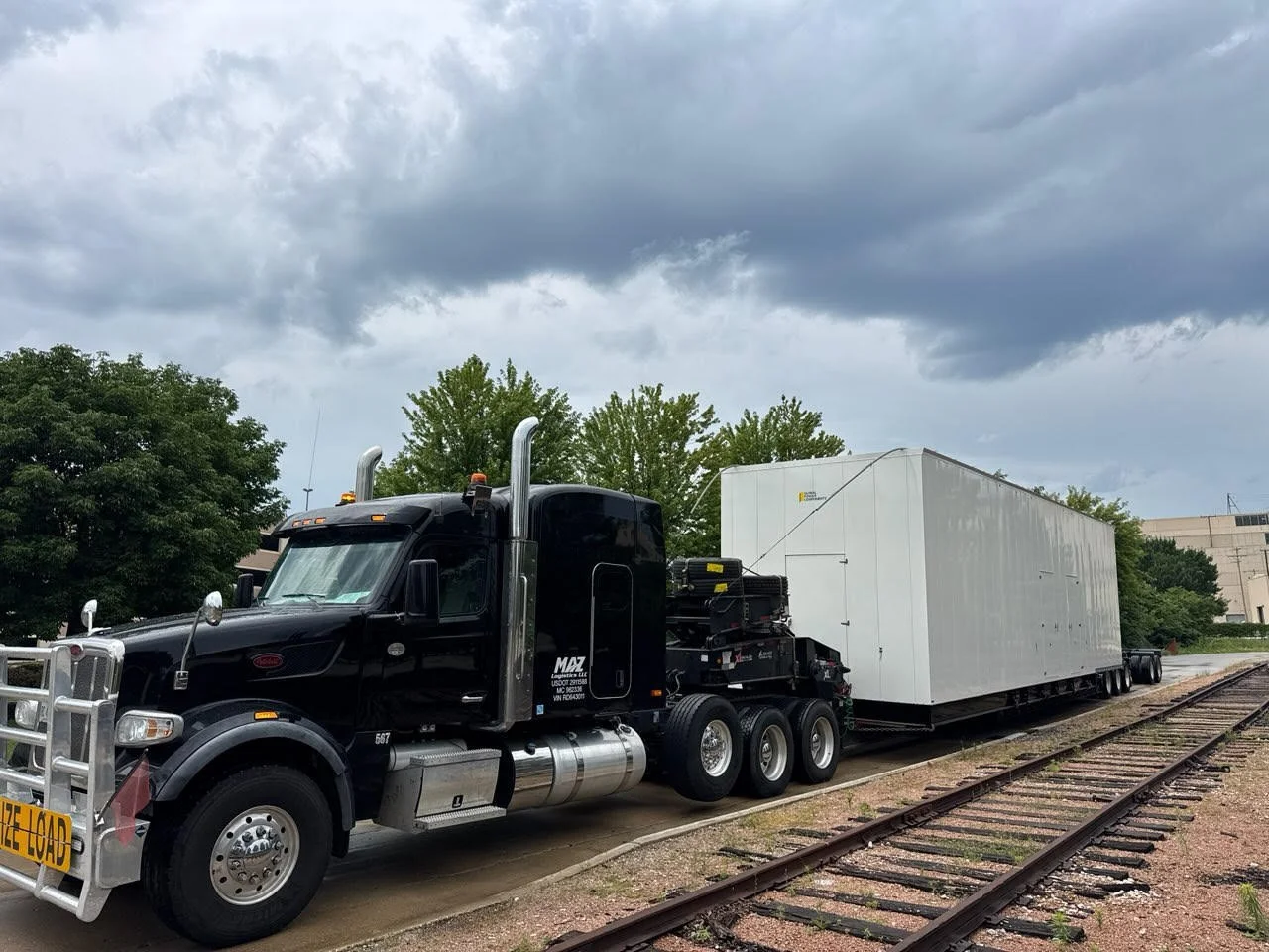 Large black semi-truck on railroad tracks carrying a massive white cargo container, under cloudy skies with green trees in the background.