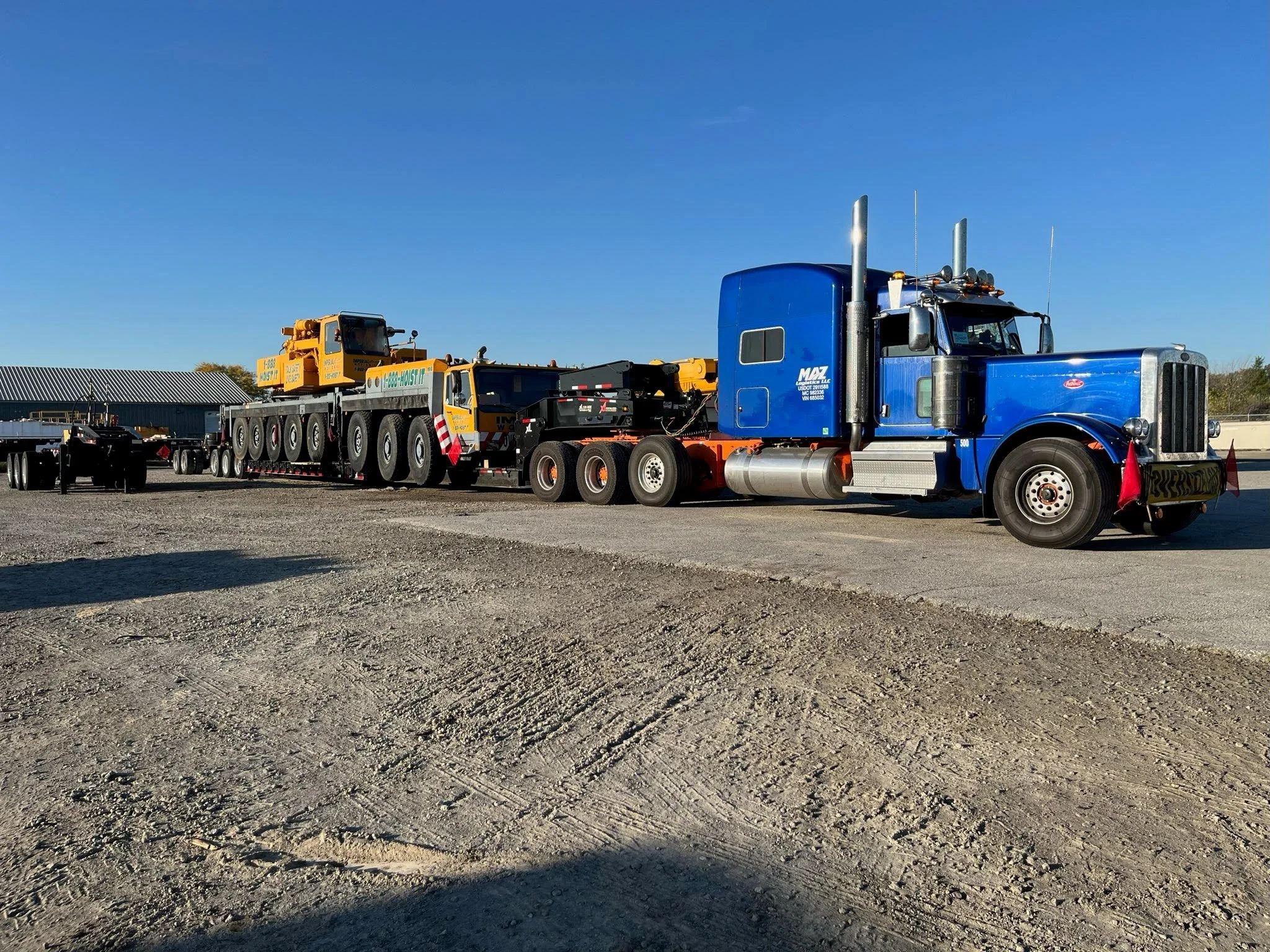 A large blue semi-truck with a trailer carrying a yellow crane on a gravel lot under a clear blue sky.