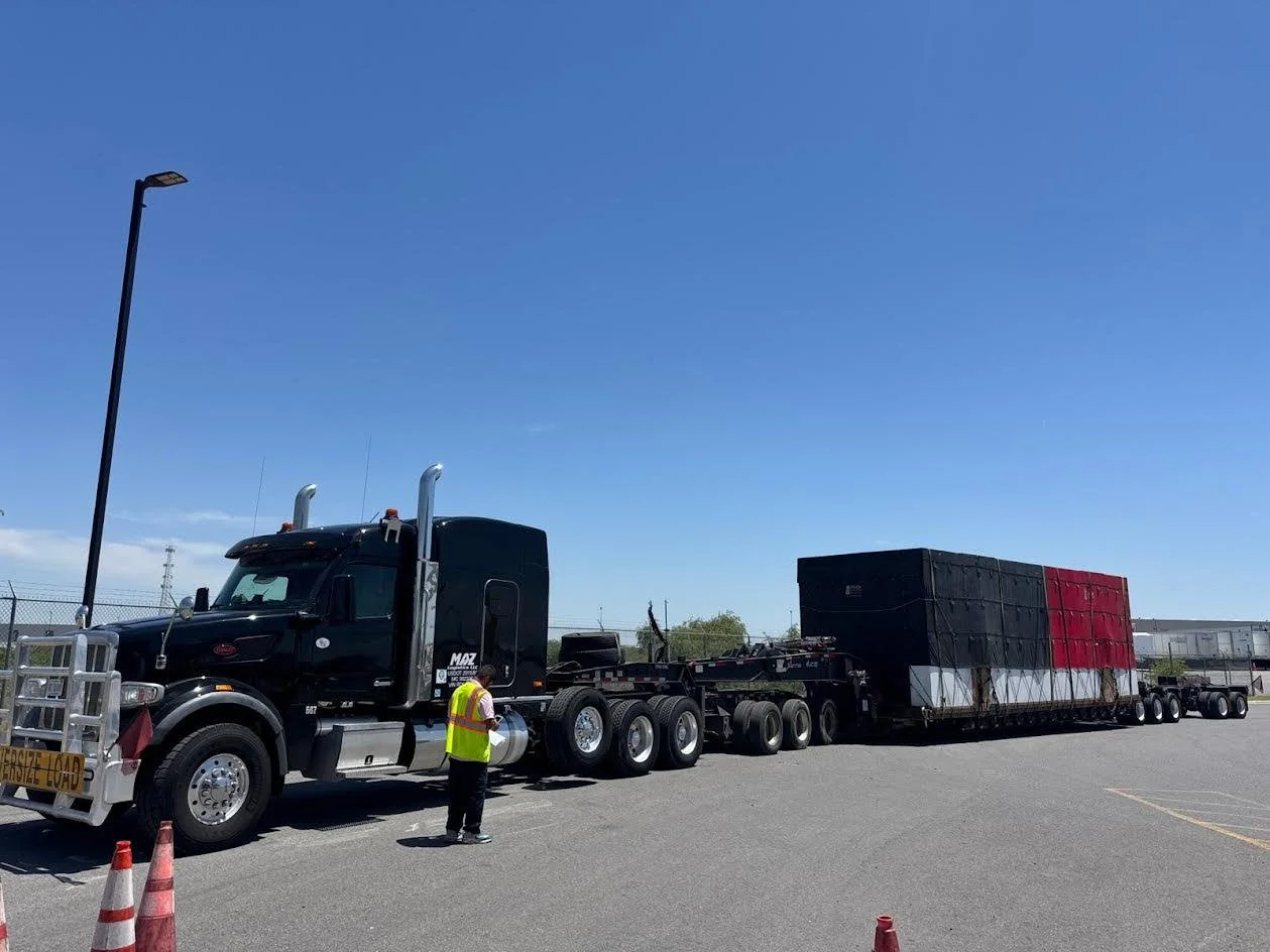 A large black semi-truck with a flatbed trailer carrying a giant rectangular object painted with a red, white, and black pattern, parked in a lot under a clear blue sky. A person in a yellow safety vest is standing next to the truck.