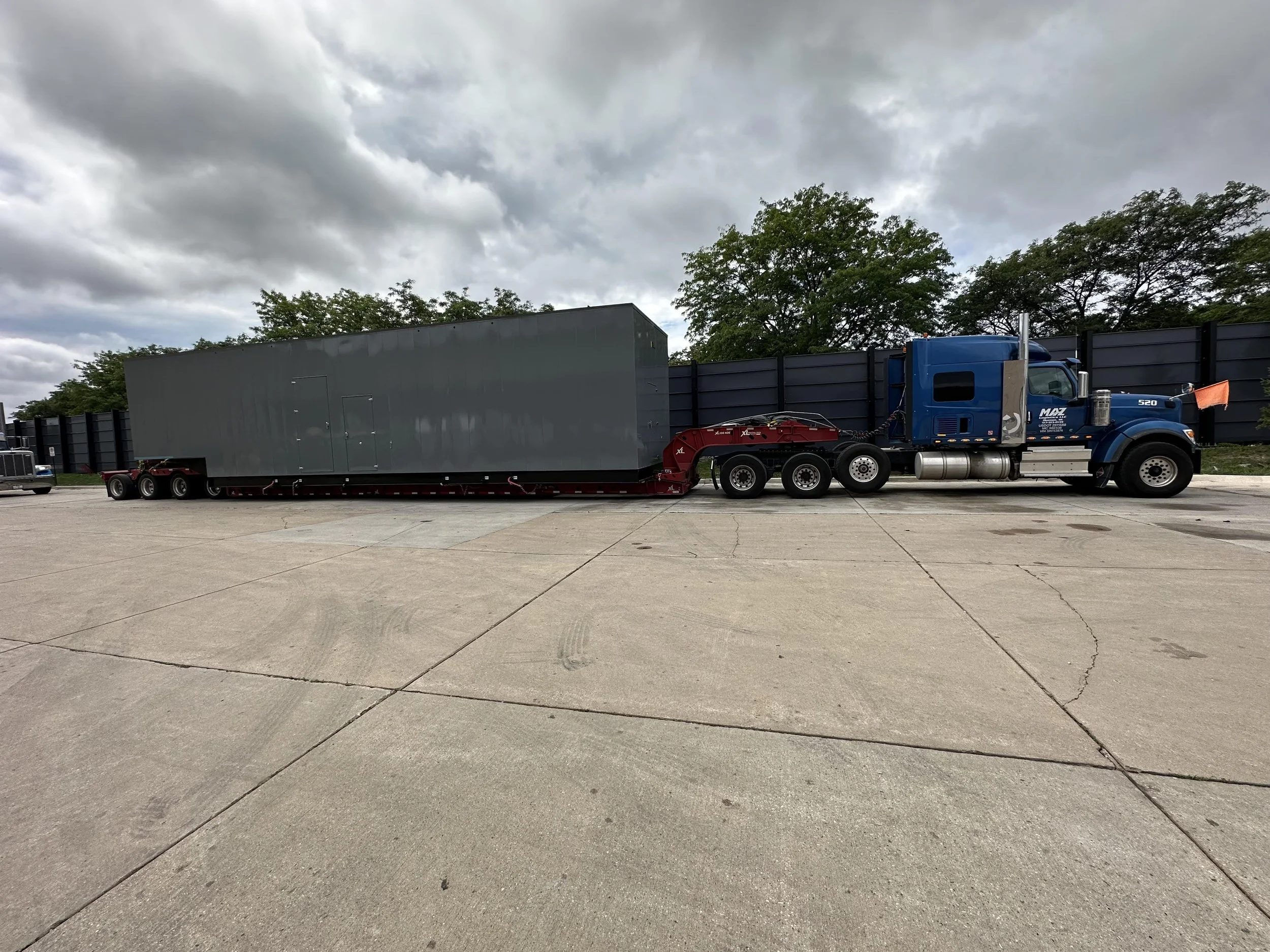 A blue semi-truck with a gray trailer parked on a wide concrete lot under cloudy skies, with trees and a black fence in the background.
