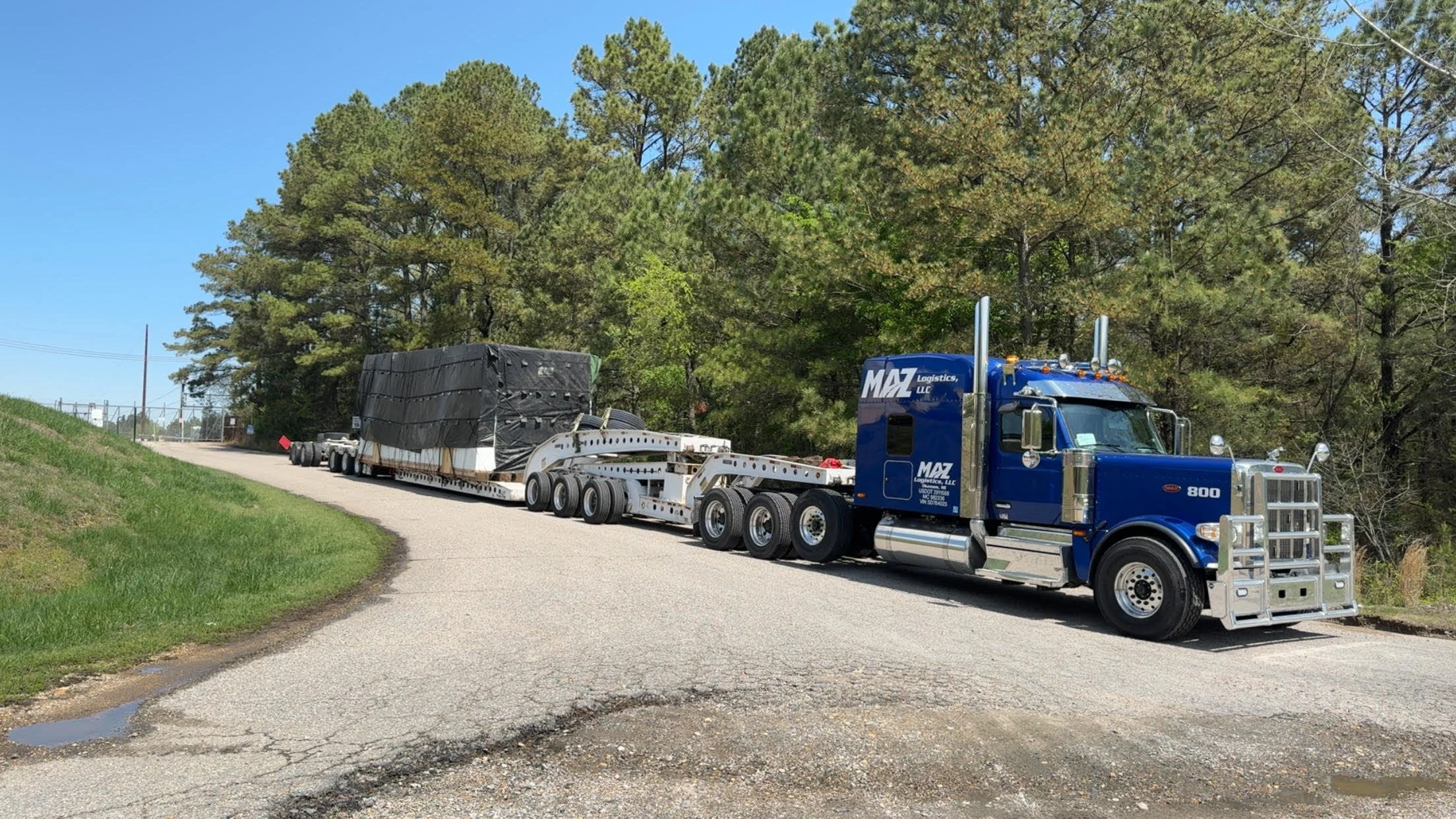 Large blue flatbed truck hauling a heavy load on a rural road, surrounded by trees and greenery.
