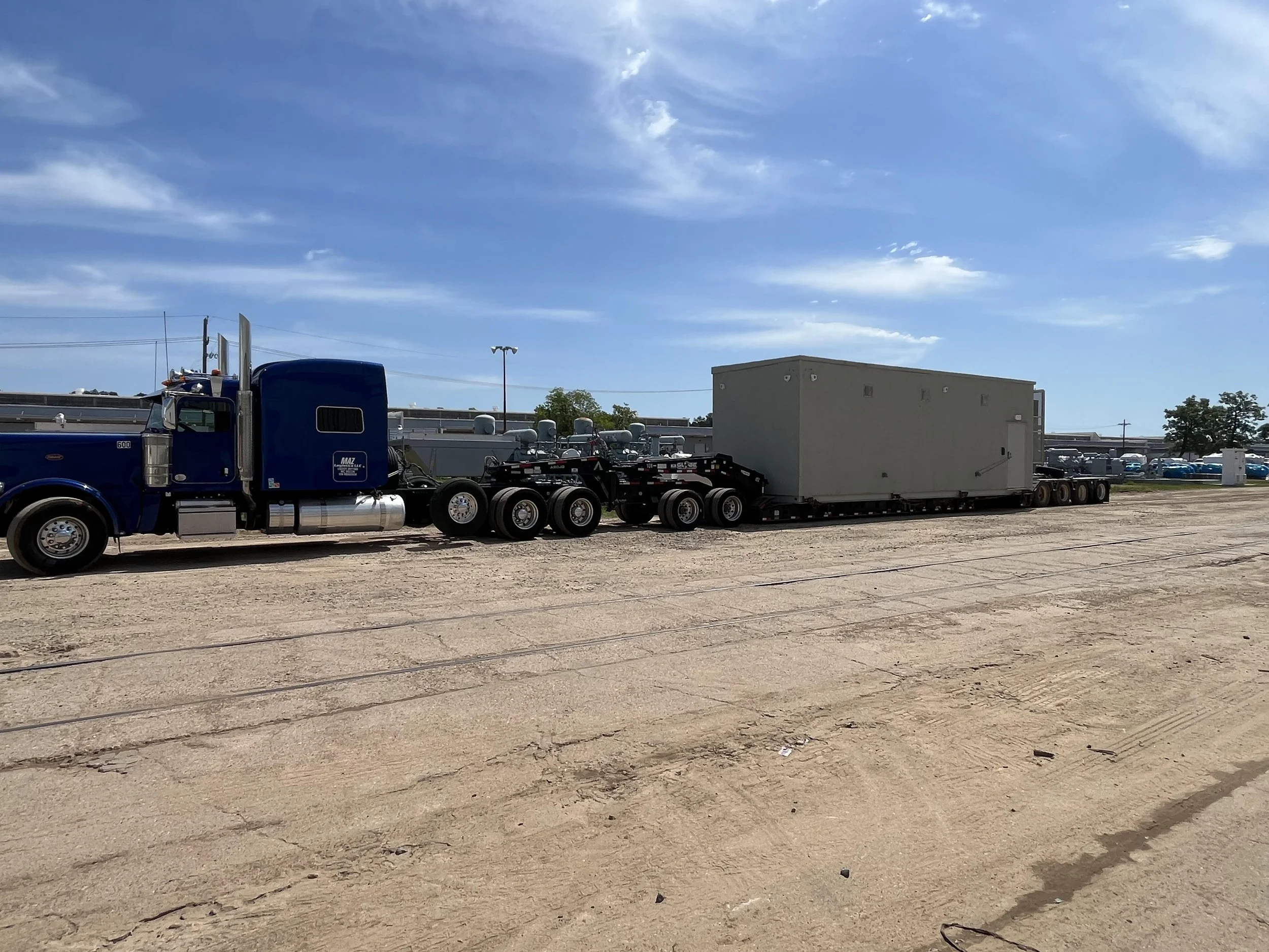 A large blue semi-truck hauling a gray modular building on a flatbed trailer on a dirt lot under a blue sky with some clouds.