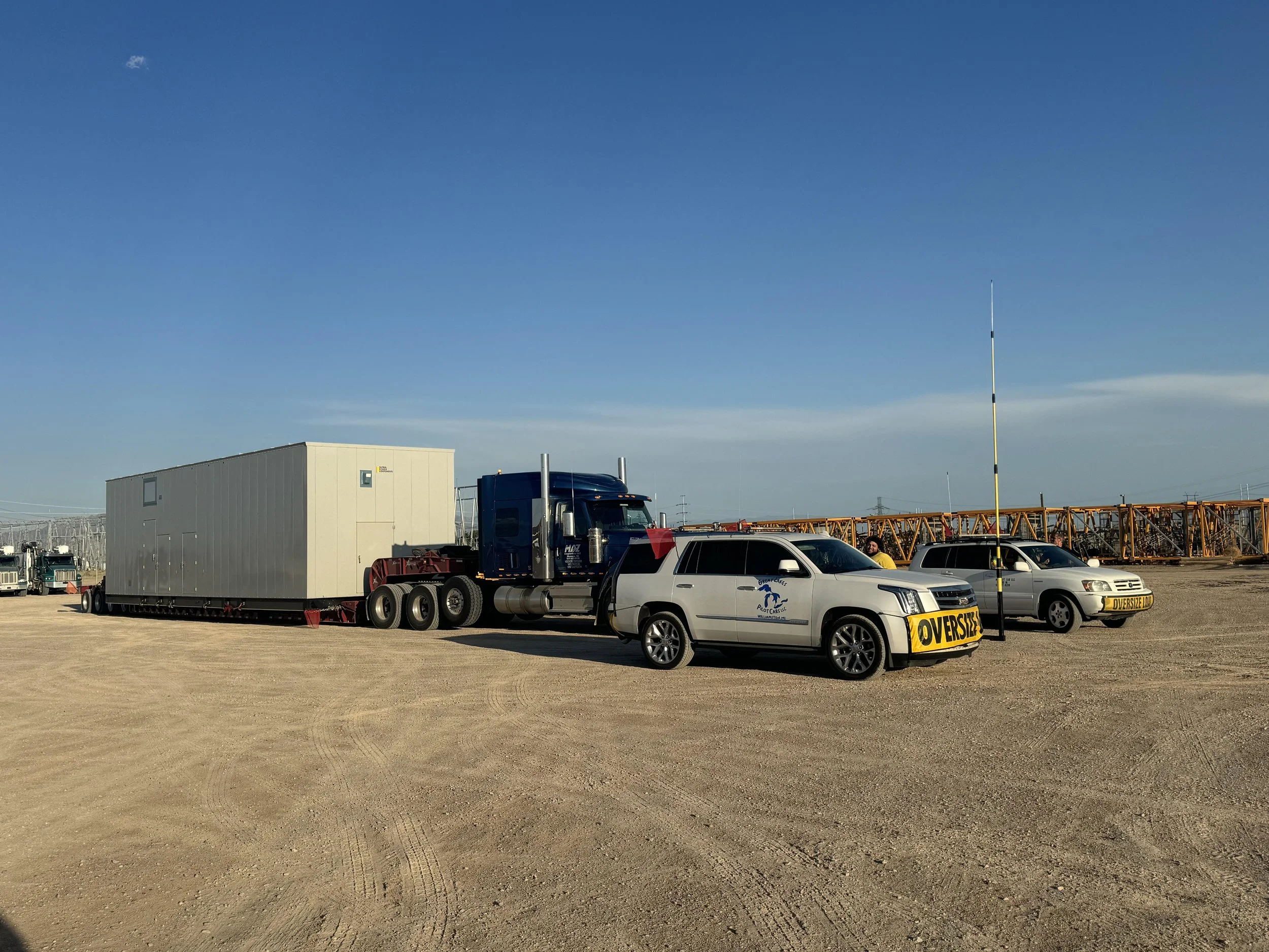 A large semi-truck with a blue cab and white trailer on a dirt lot, accompanied by two silver cars with yellow 'Oversize Load' signs, one with a logo of a dolphin and the text 'Sealife Molokai LLC,' and a person standing near the second car, with a l
