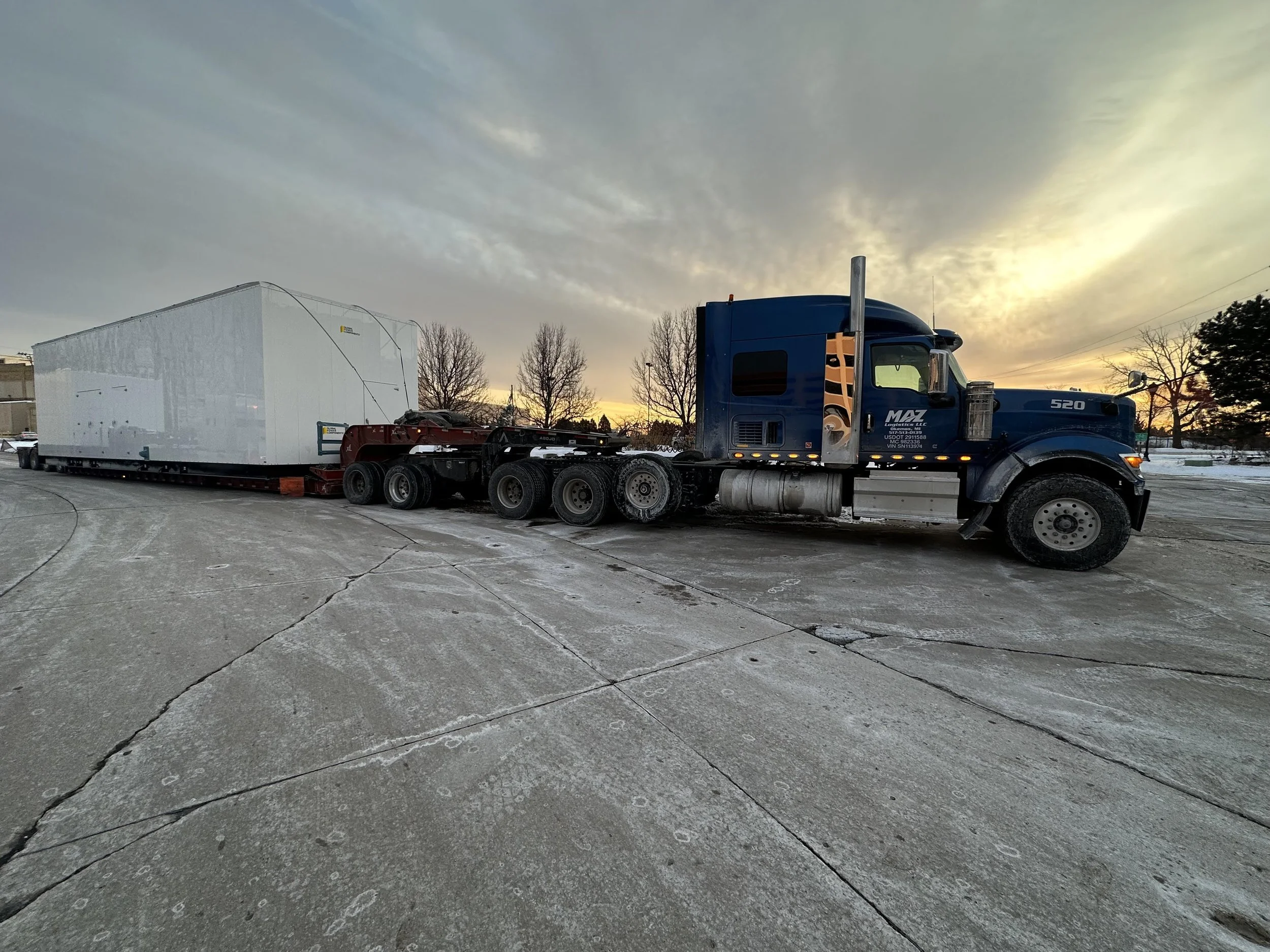 A large blue semi-truck parked on a concrete lot with a white enclosed trailer attached beneath a cloudy sky at sunset.