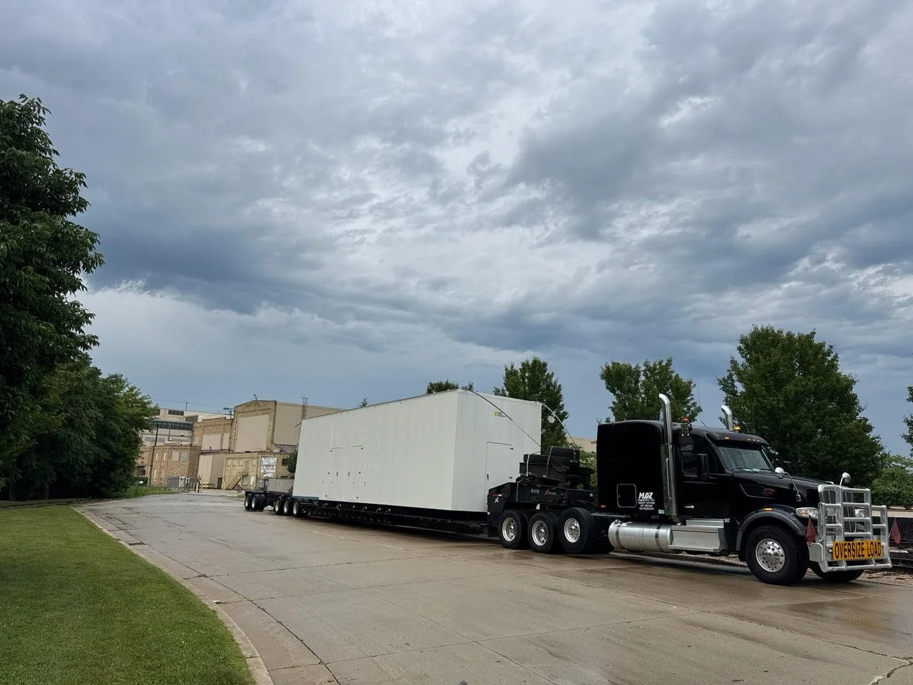 A large black semi-truck with an oversized load sign on the front, hauling a big white rectangular object on a flatbed trailer on a wet road under a cloudy sky, with trees on the side and industrial buildings in the background.