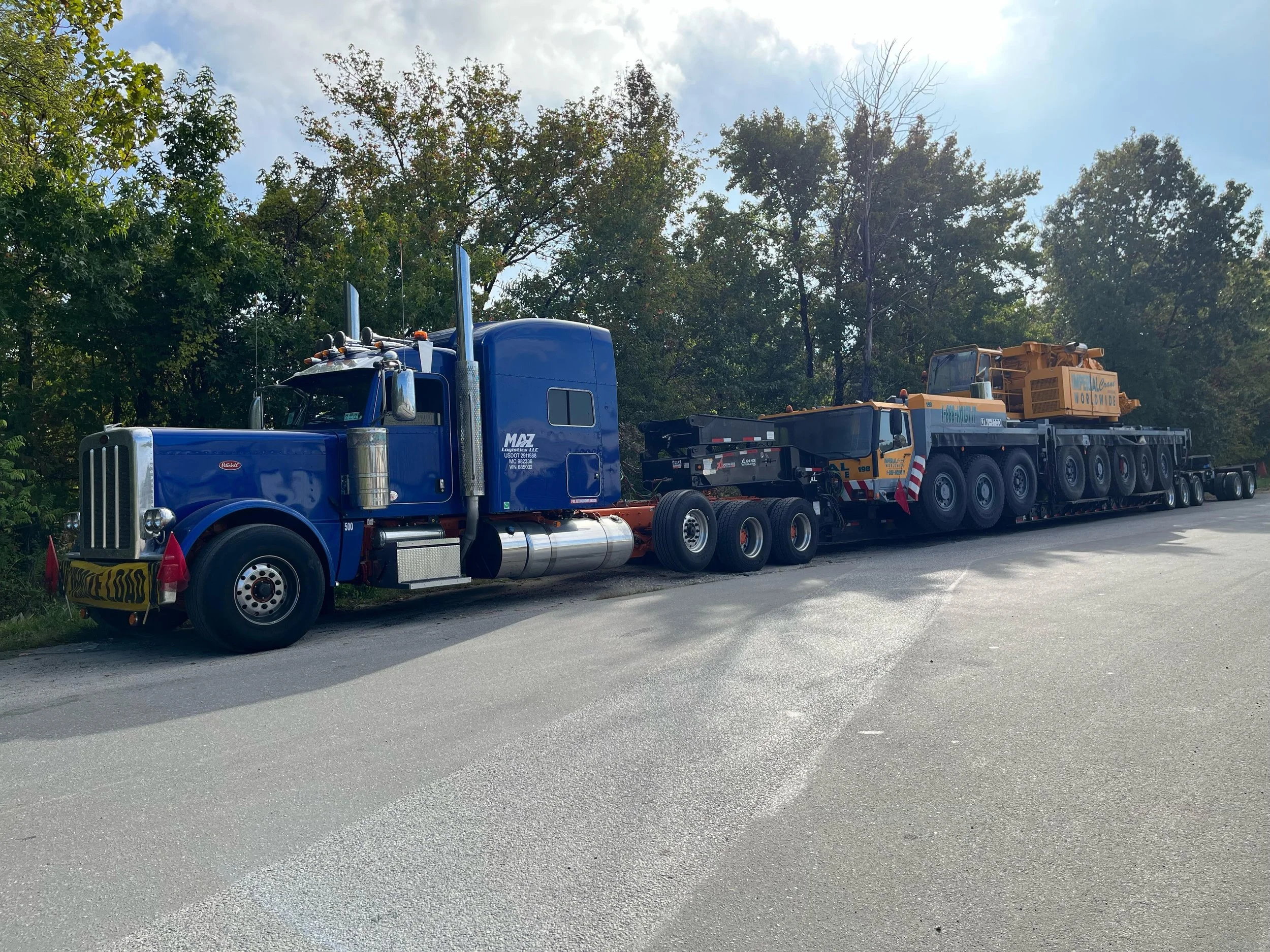 A large blue semi-truck with a flatbed trailer carrying a yellow crane, parked on the side of a road with trees in the background.