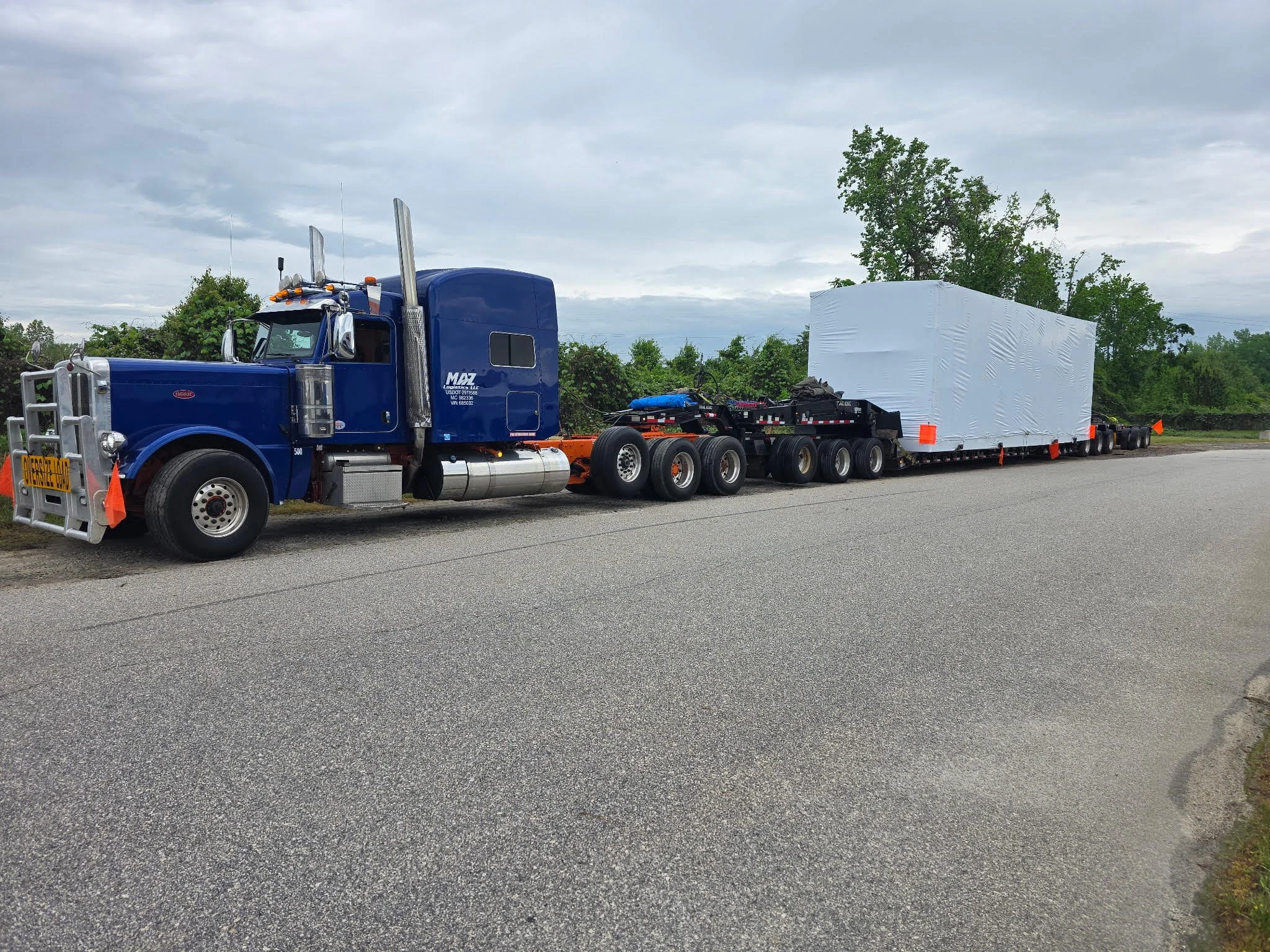 A large blue semi-truck with a white load parked on the side of a paved road on a cloudy day, with trees and green foliage in the background.