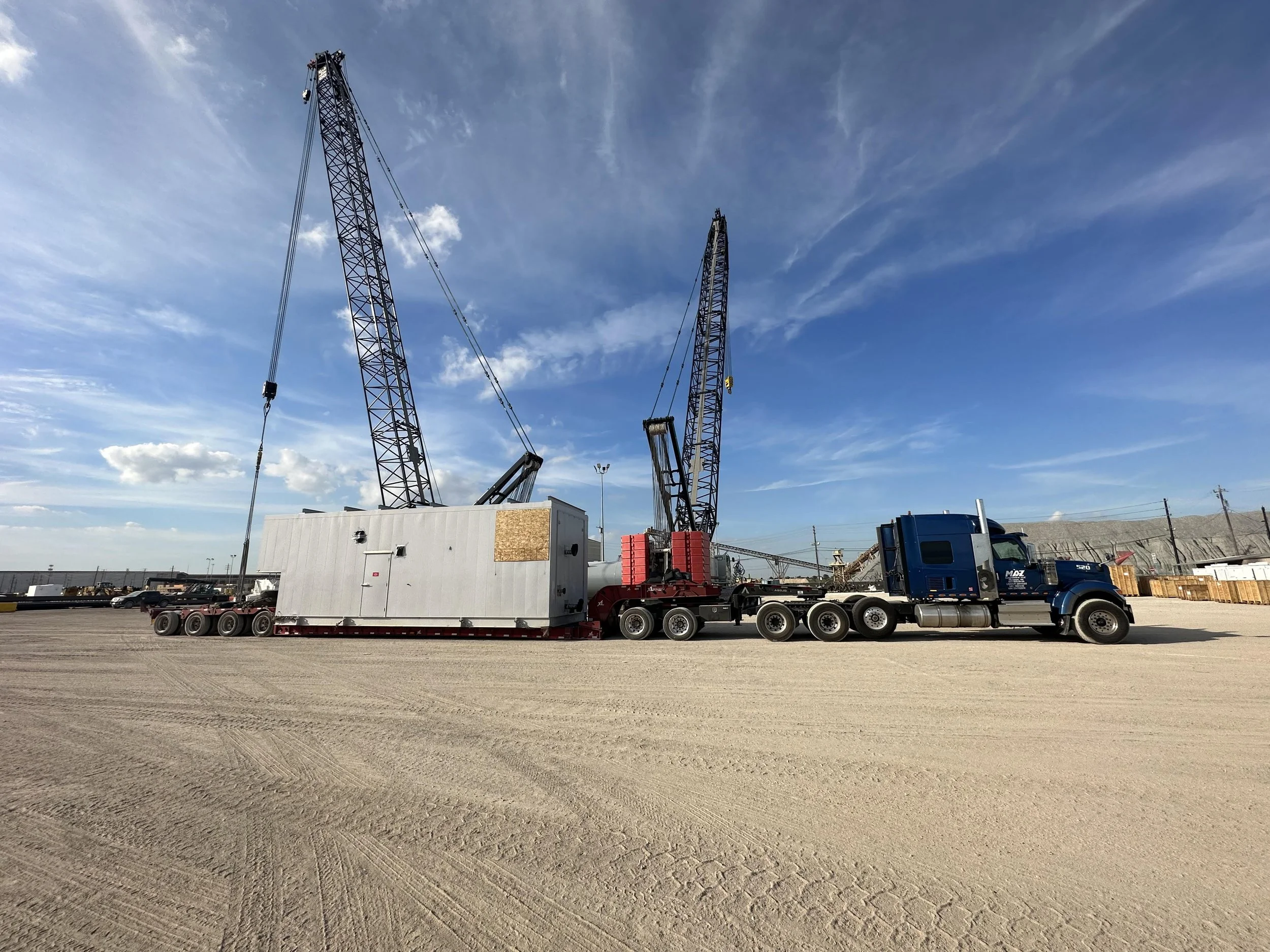 A large semi-truck with a trailer carrying two tall crane booms parked on a dirt lot under a blue sky with scattered clouds.