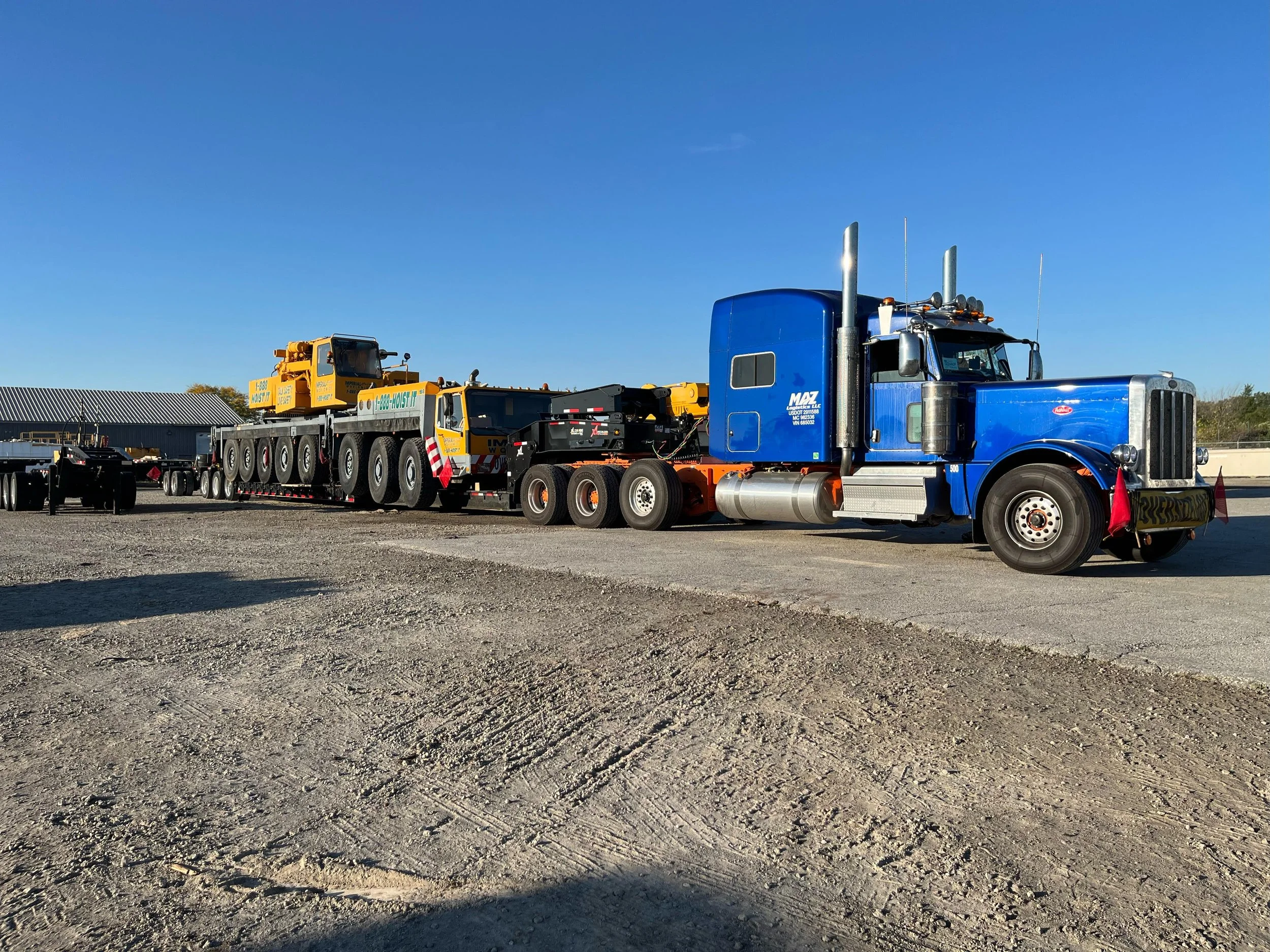 Blue semi-truck with a large yellow crane on a flatbed trailer parked on a gravel lot on a clear day.