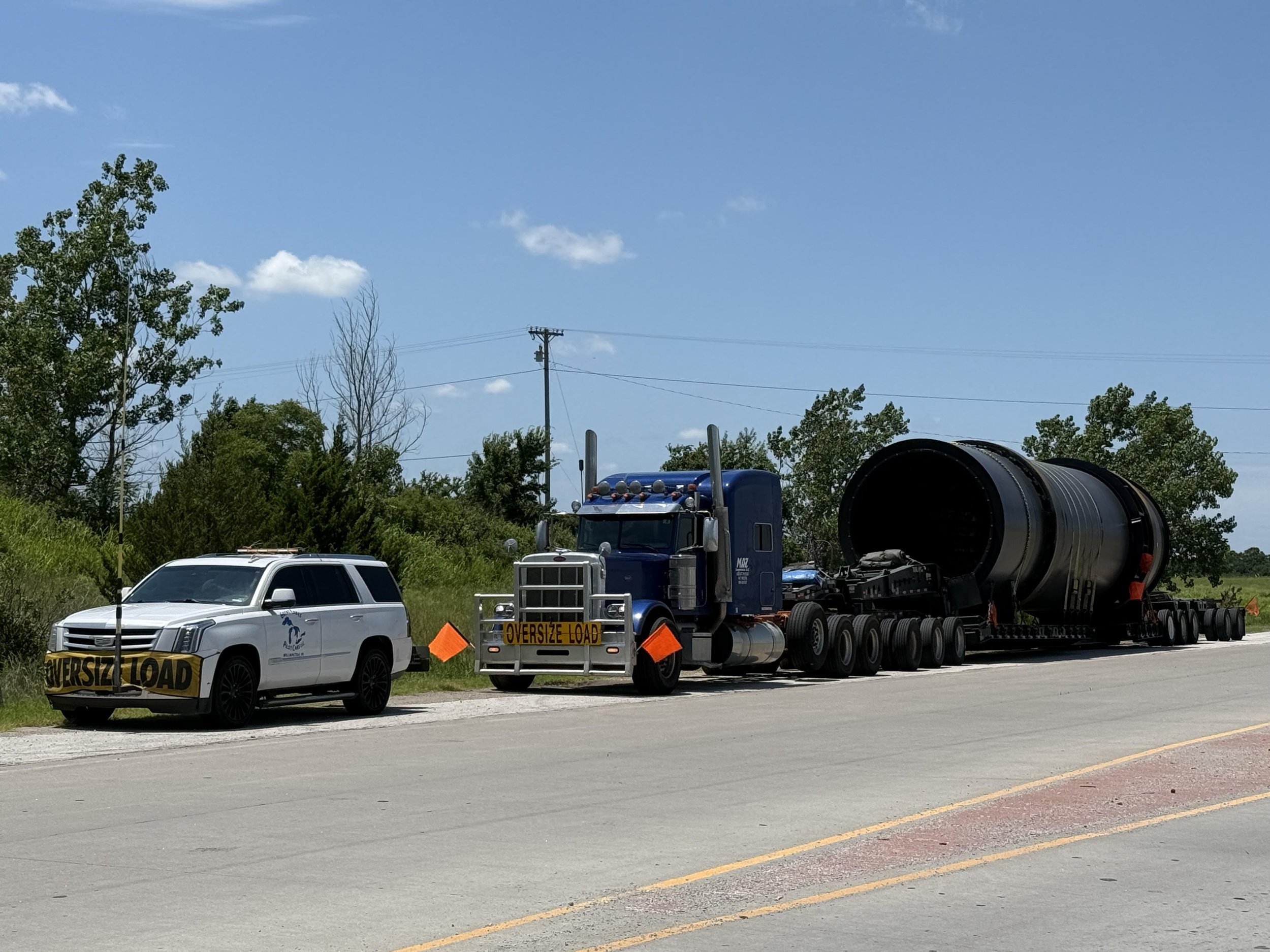 A white SUV and a large blue semi-truck on the side of the road, both with 'Oversize Load' signs. The semi-truck is hauling a very large cylindrical object. The background includes trees and utility poles under a partly cloudy sky.