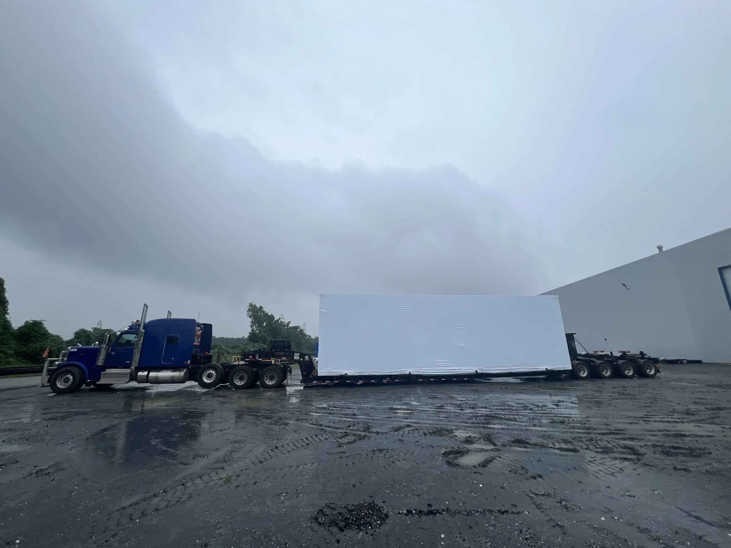 Semi-truck transporting a large, rectangular, white object on a wet, muddy ground under cloudy sky.