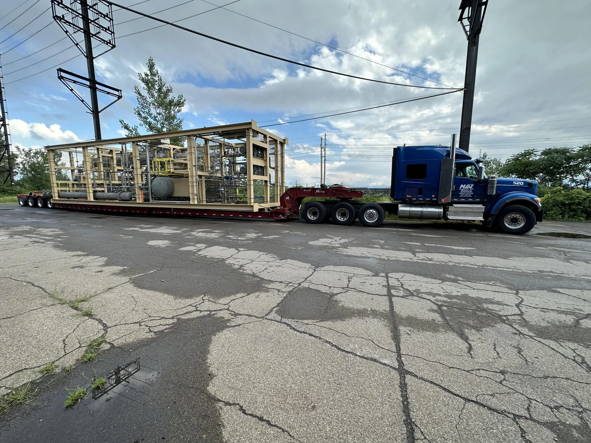 A large truck transporting industrial equipment with a complex network of pipes and tanks on a flatbed trailer, parked on a cracked asphalt road under cloudy skies.