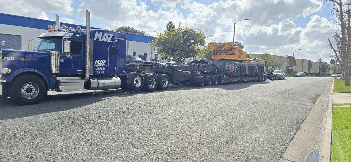 A large blue semi-truck with a flatbed trailer transporting heavy equipment parked on a city street.