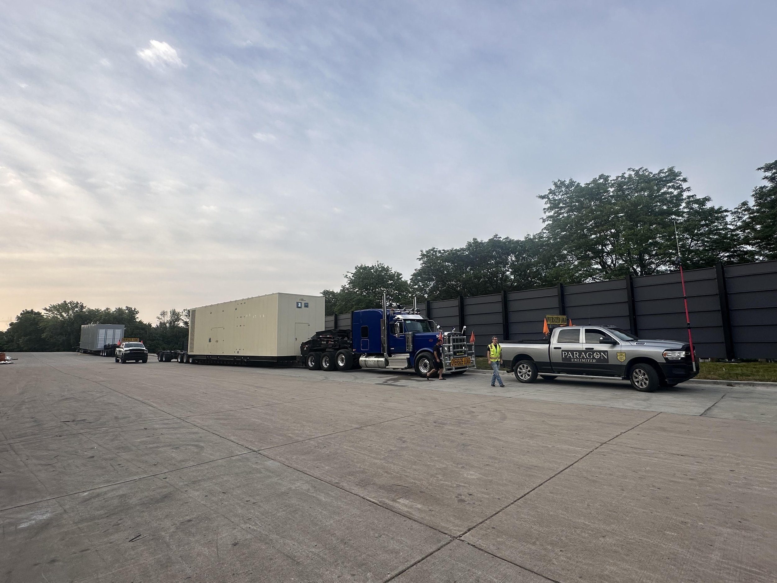 Large truck with a blue cab and black trailer, carrying a large white container, parked on a concrete lot with other vehicles and people nearby, against a backdrop of trees and a gray fence.