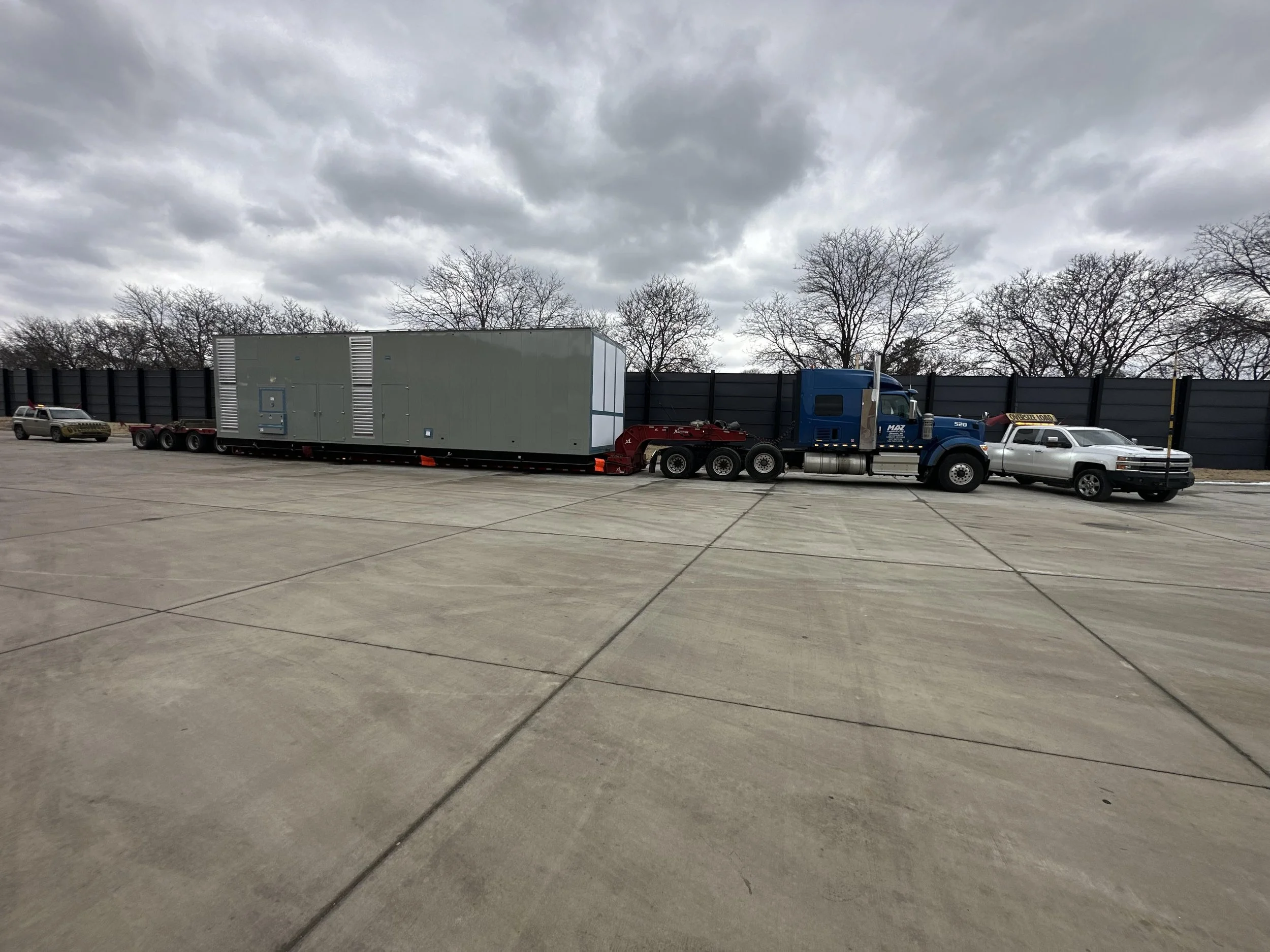 A large semi-truck with a blue cab and gray trailer transporting a large gray generator, parked on an outdoor concrete lot under cloudy skies. There are other trucks and vehicles nearby, with a black fence and leafless trees in the background.