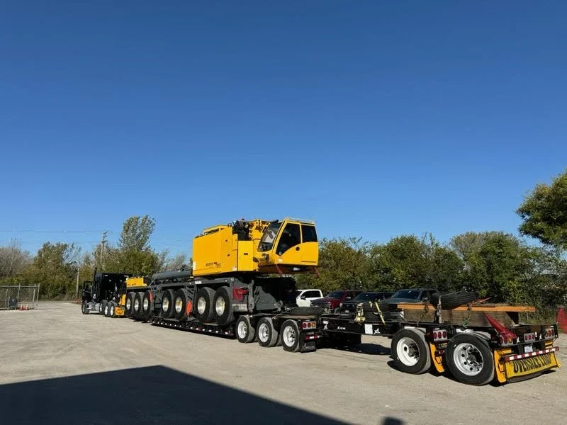 Large flatbed truck transporting a yellow construction crane on a clear day with a blue sky and trees in the background.