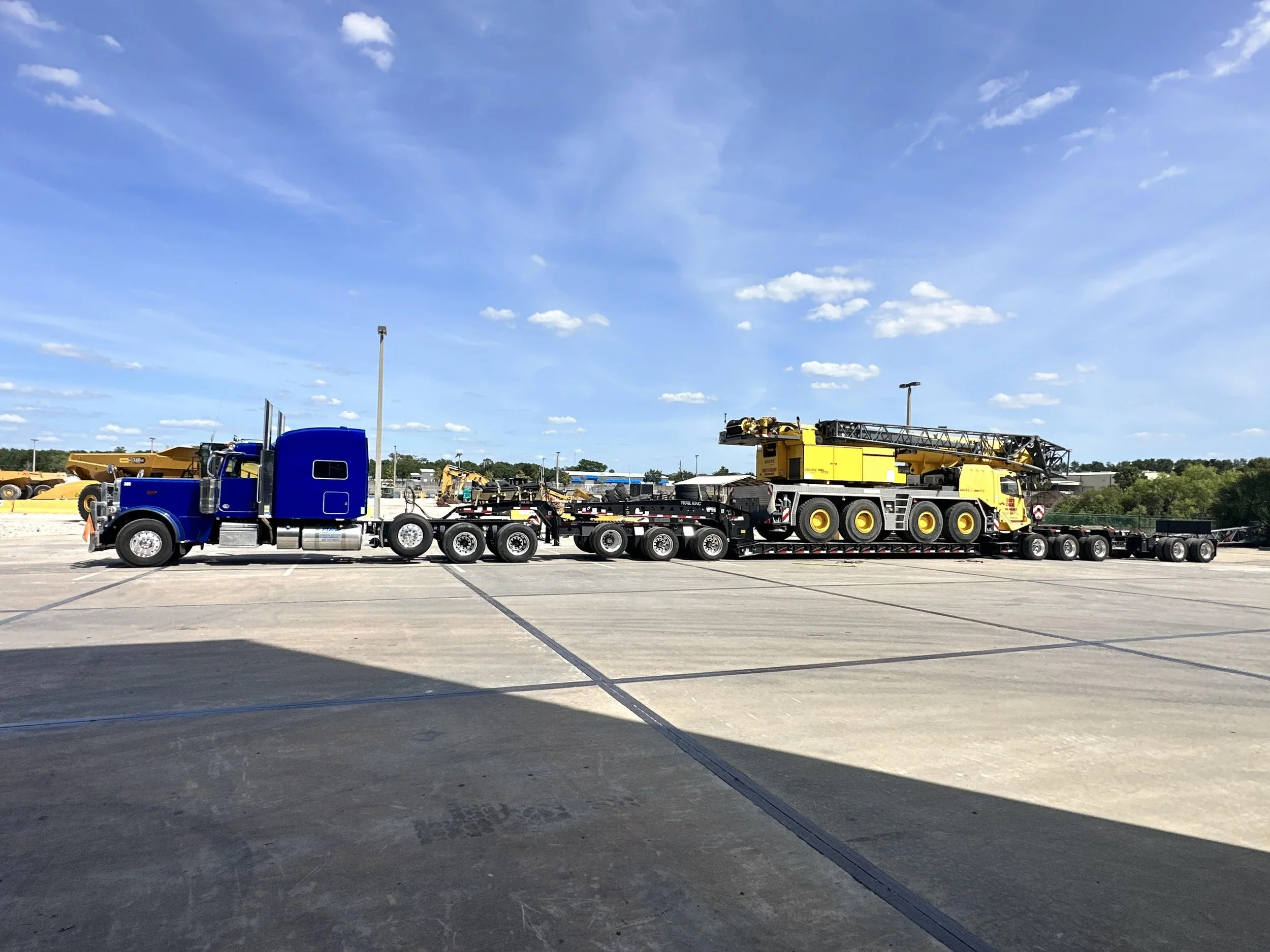 A large blue semi-truck with a yellow crane on a lowboy trailer in a parking lot on a clear, sunny day.
