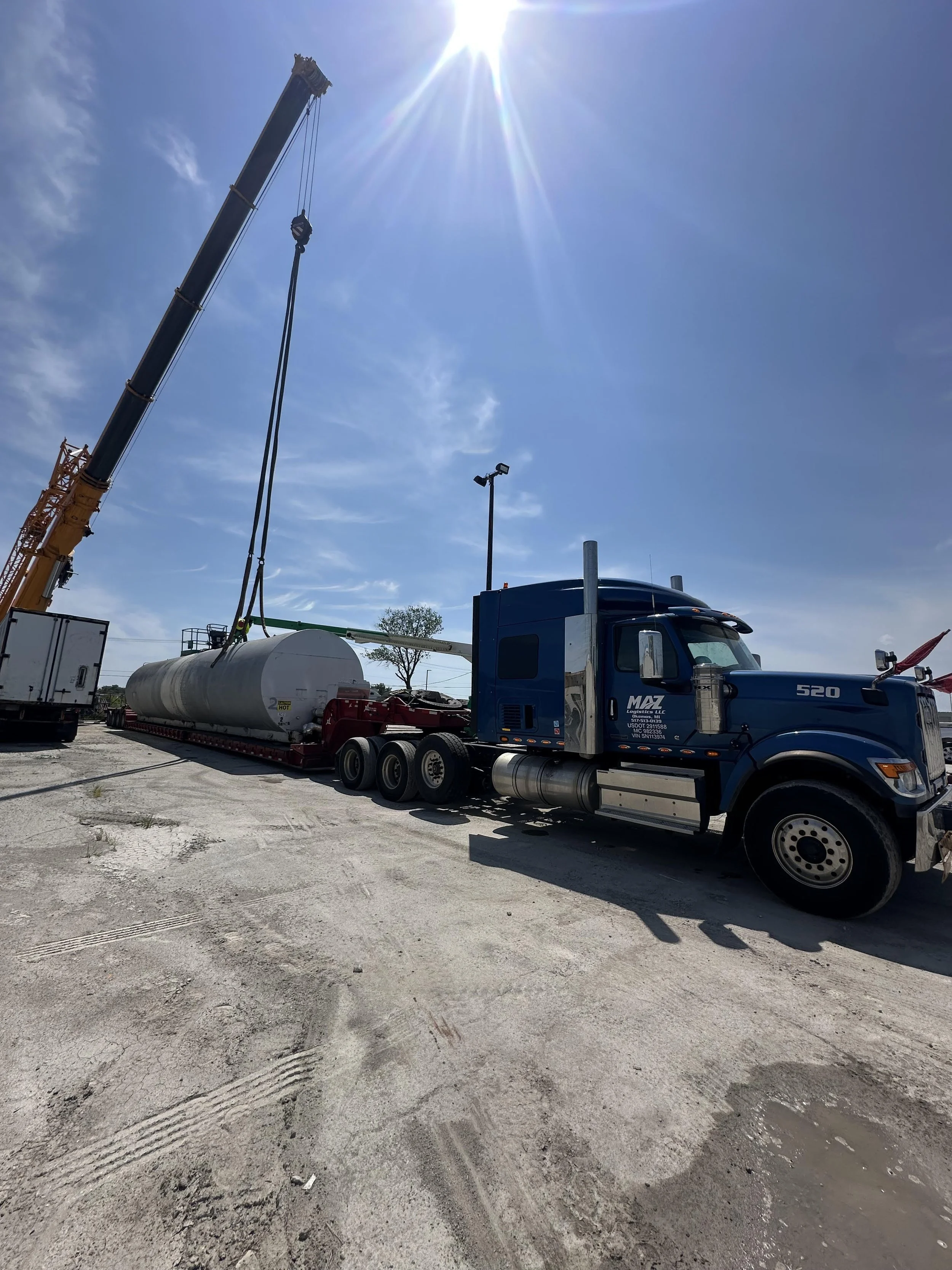 A large blue semi-truck with a tank trailer being lifted by a crane on a sunny day with a clear sky.