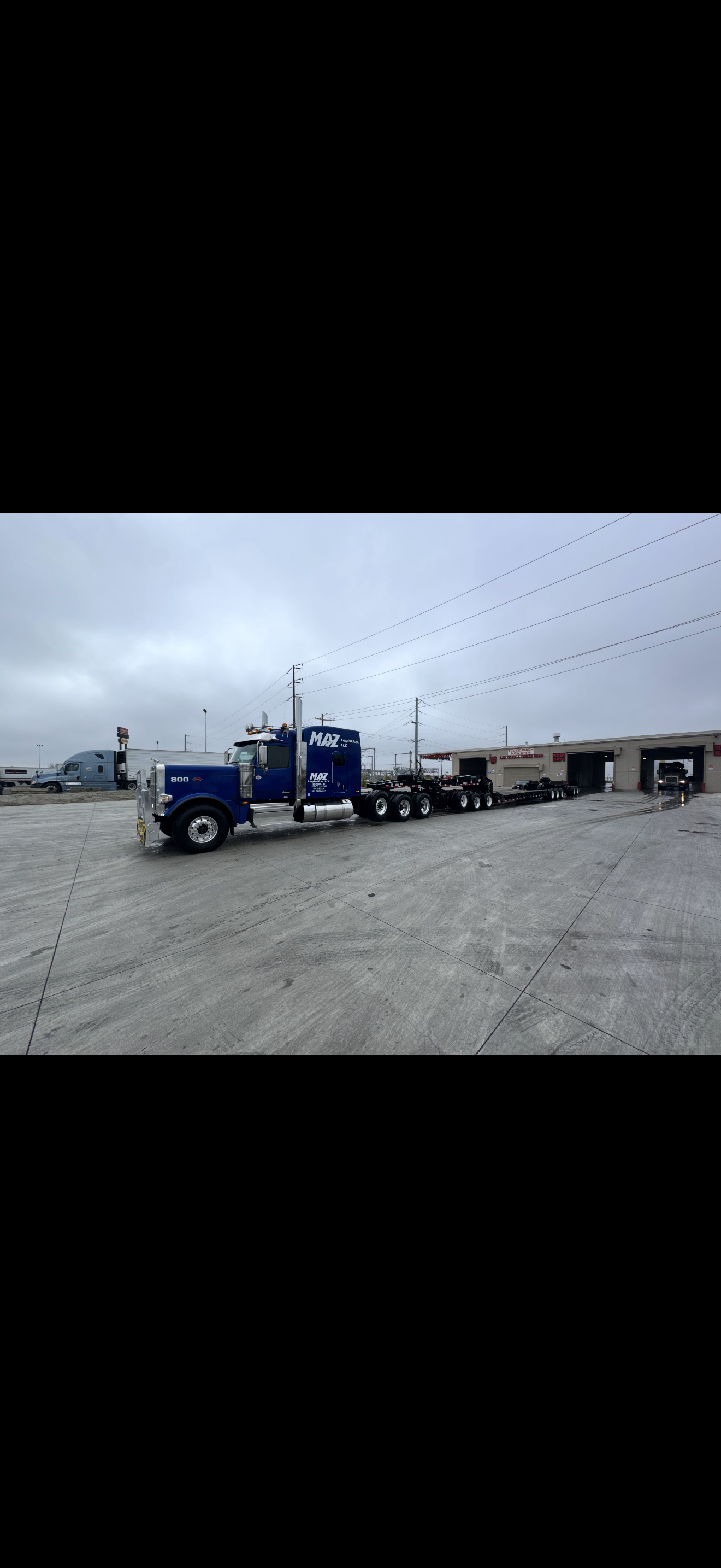 Blue semi-truck with a flatbed trailer parked outside a building under cloudy sky.