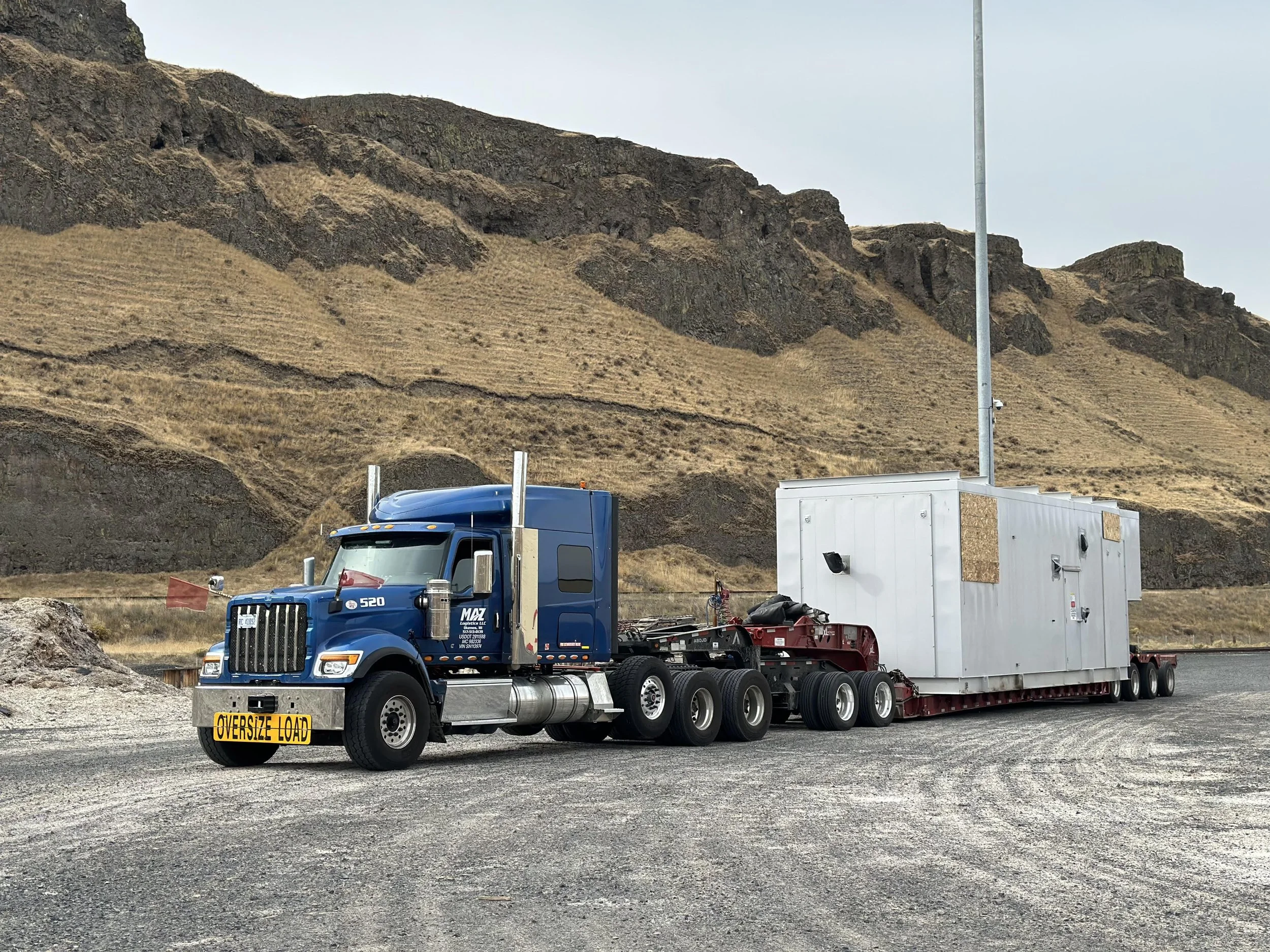 Large blue semi-truck parked on gravel ground in front of a rocky hillside, carrying a white container on extended trailer, with a sign that reads 'OVERSIZE LOAD' on the front of the truck.