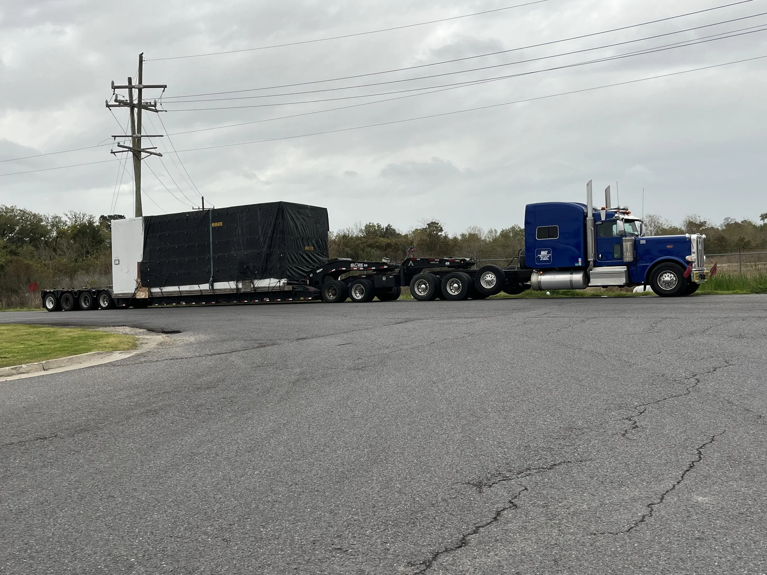 A blue semi-truck with a flatbed trailer transporting a large object covered with black tarp on a paved road under cloudy sky.
