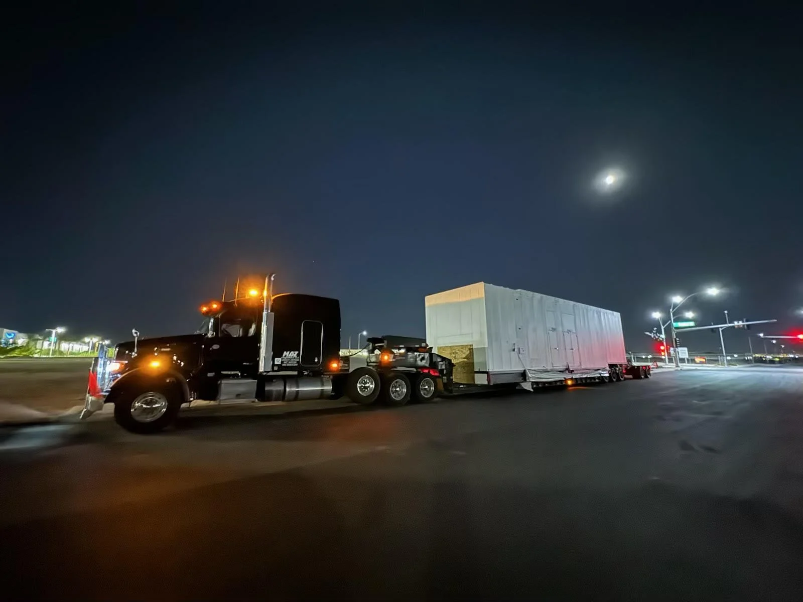 Nighttime scene of a large black semi-truck with a white trailer driving on a city street, illuminated by streetlights, with a visible moon in the night sky.