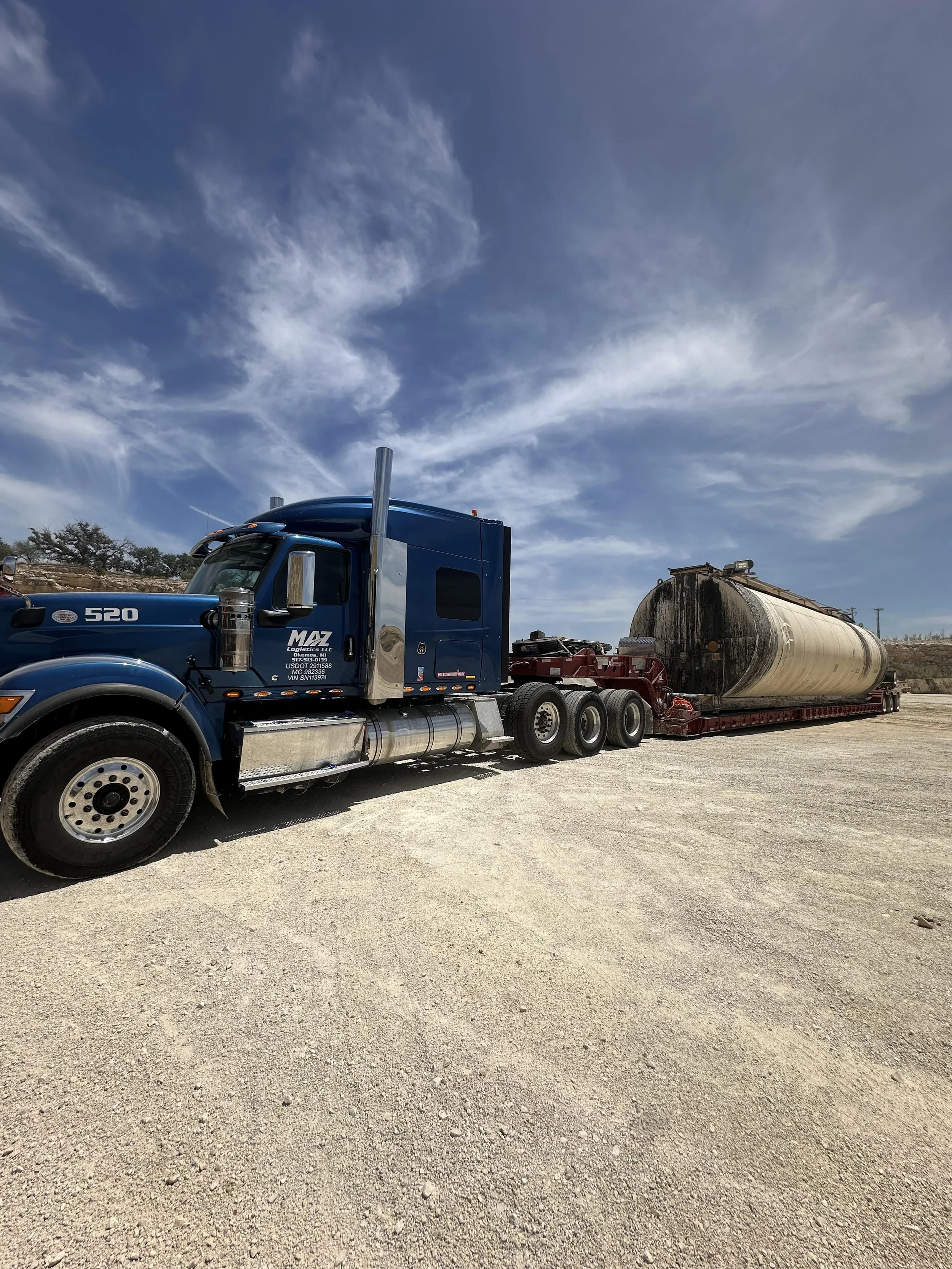 A blue semi-truck with a tanker trailer on a gravel lot under a partly cloudy sky.