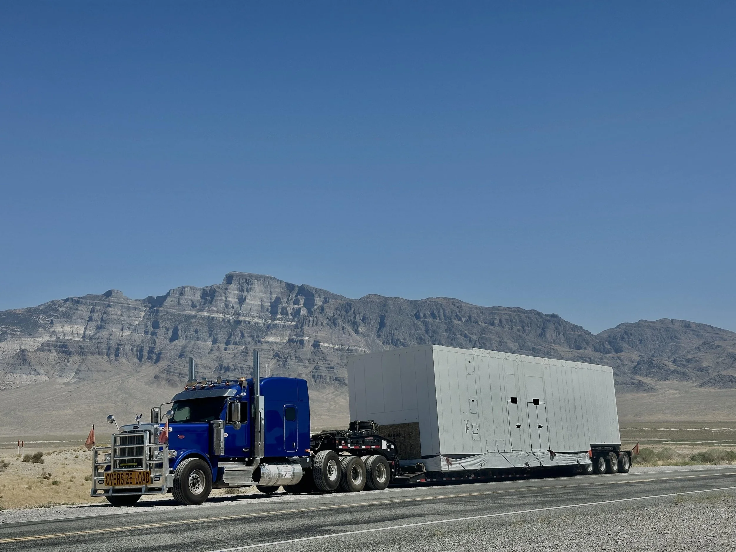 A blue semi-truck hauling a large white trailer on a highway with mountainous desert landscape in the background.