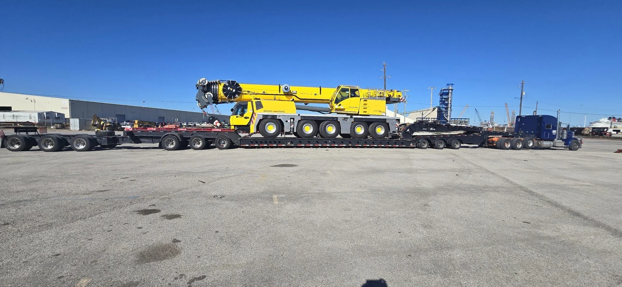 A large yellow and gray crane on a flatbed trailer in an industrial area or port, with clear blue sky above.