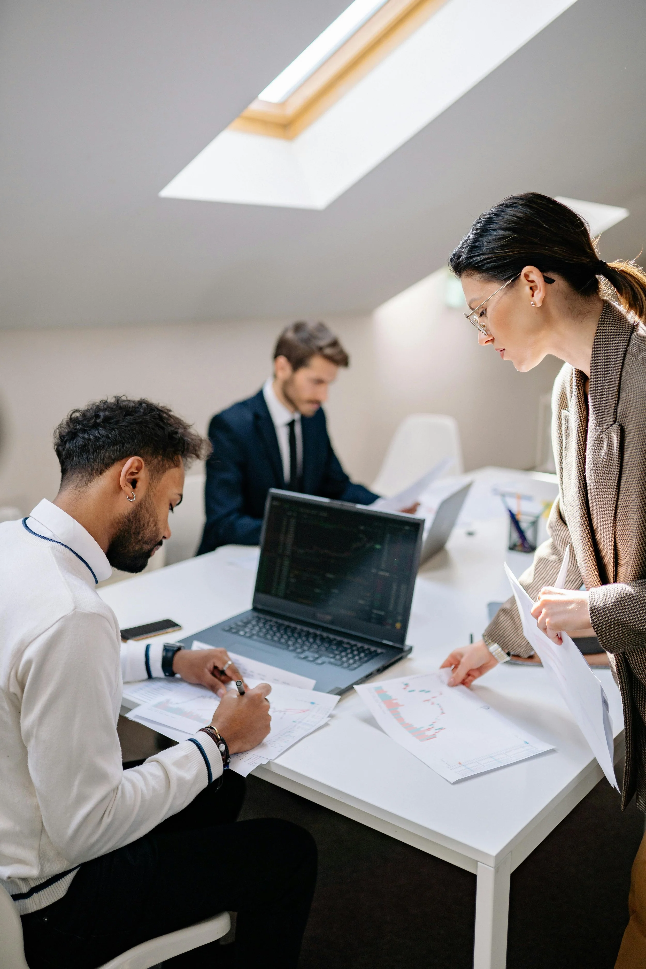 A business meeting with three professionals reviewing documents and working on laptops in a conference room with skylights.