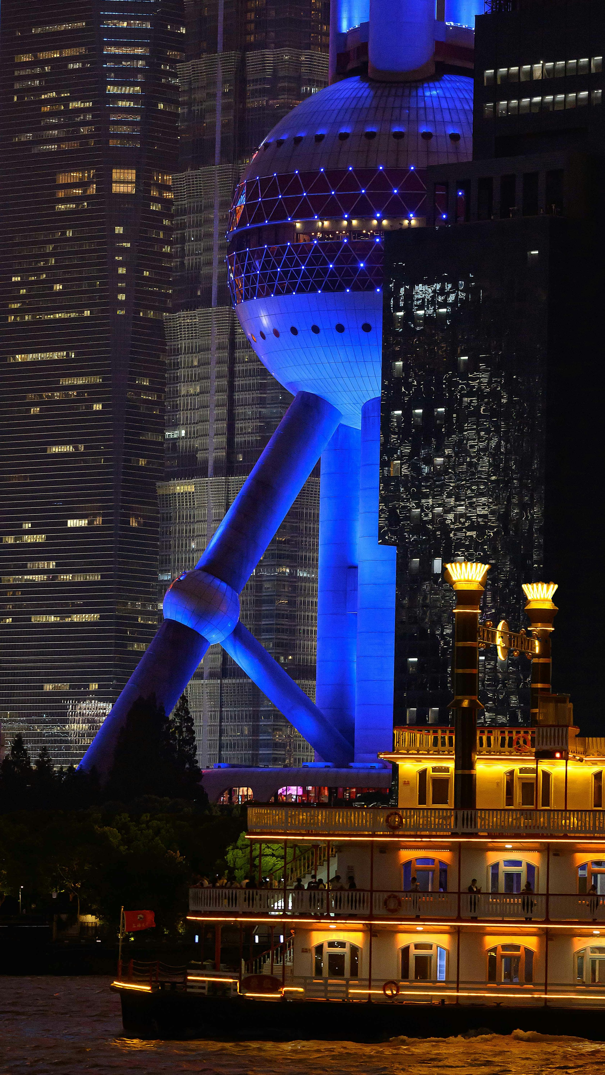 Night view of the Shanghai Tower, with its illuminated spire and observation deck, towering over illuminated modern skyscrapers; in the foreground, a brightly lit vintage-style river cruise boat on the water.