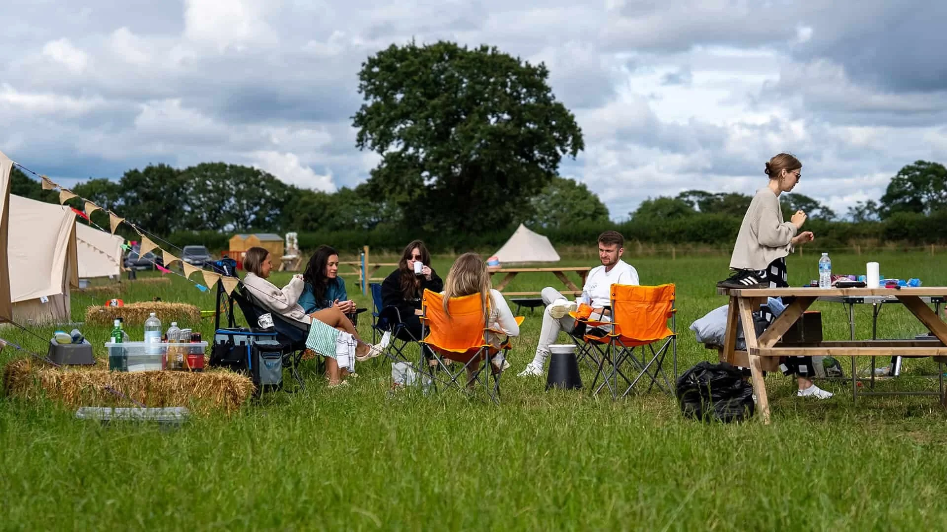 A group of people sitting on camping chairs and hay bales in a grassy field during daytime, with tents and trees in the background, having a outdoor gathering or picnic.