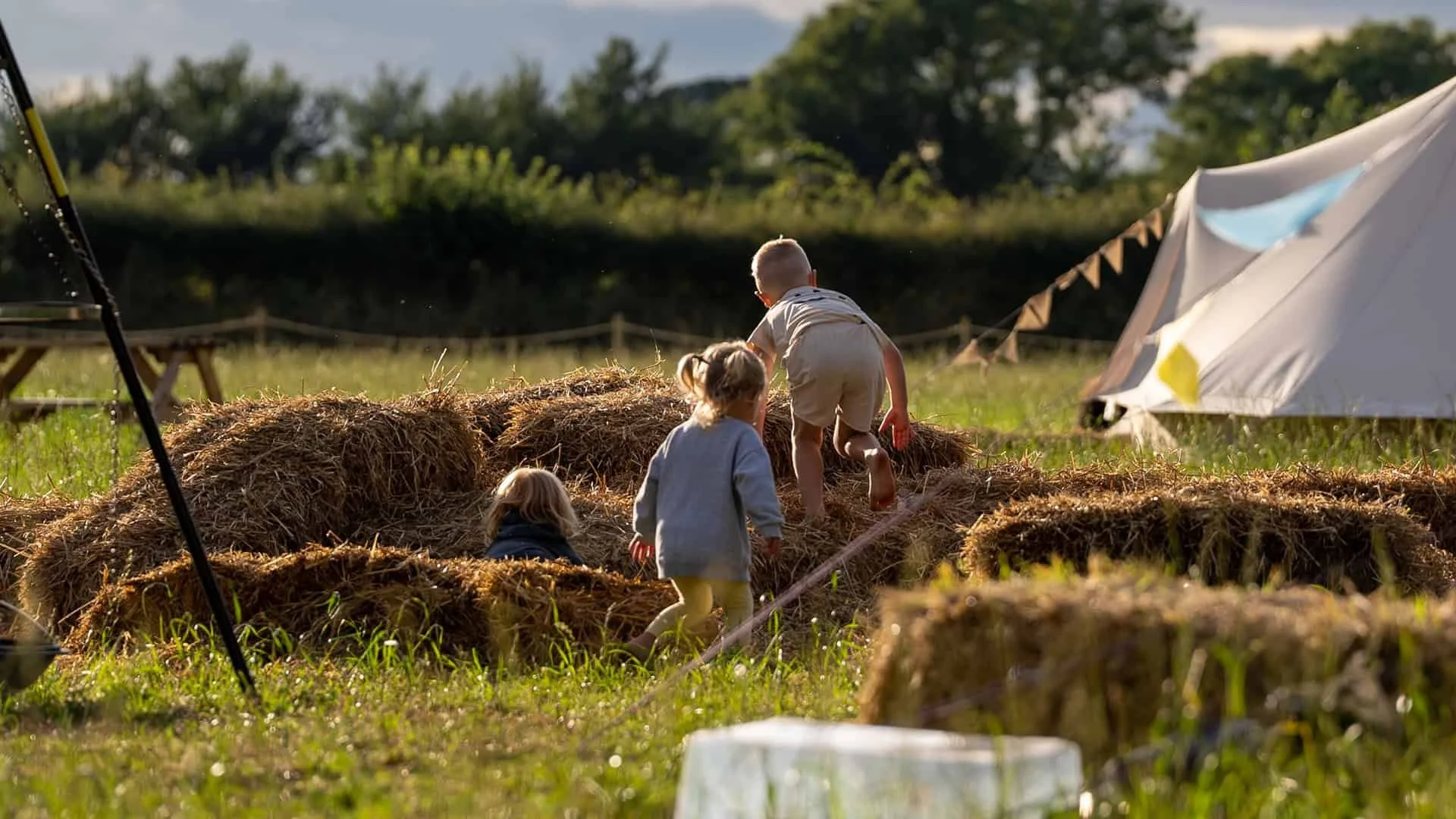 Three children playing on hay bales in a grassy field near a tent, with trees and a sunset sky in the background.