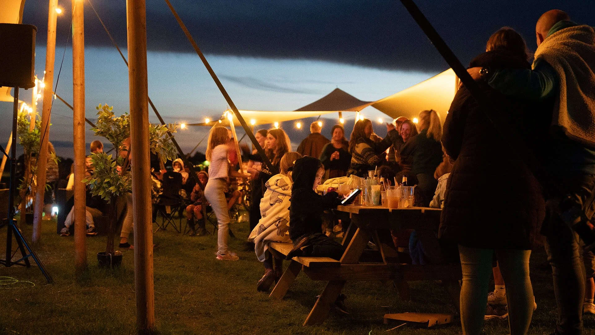 People gathered under a large canopy with string lights at night, socializing and sitting at picnic tables with drinks.