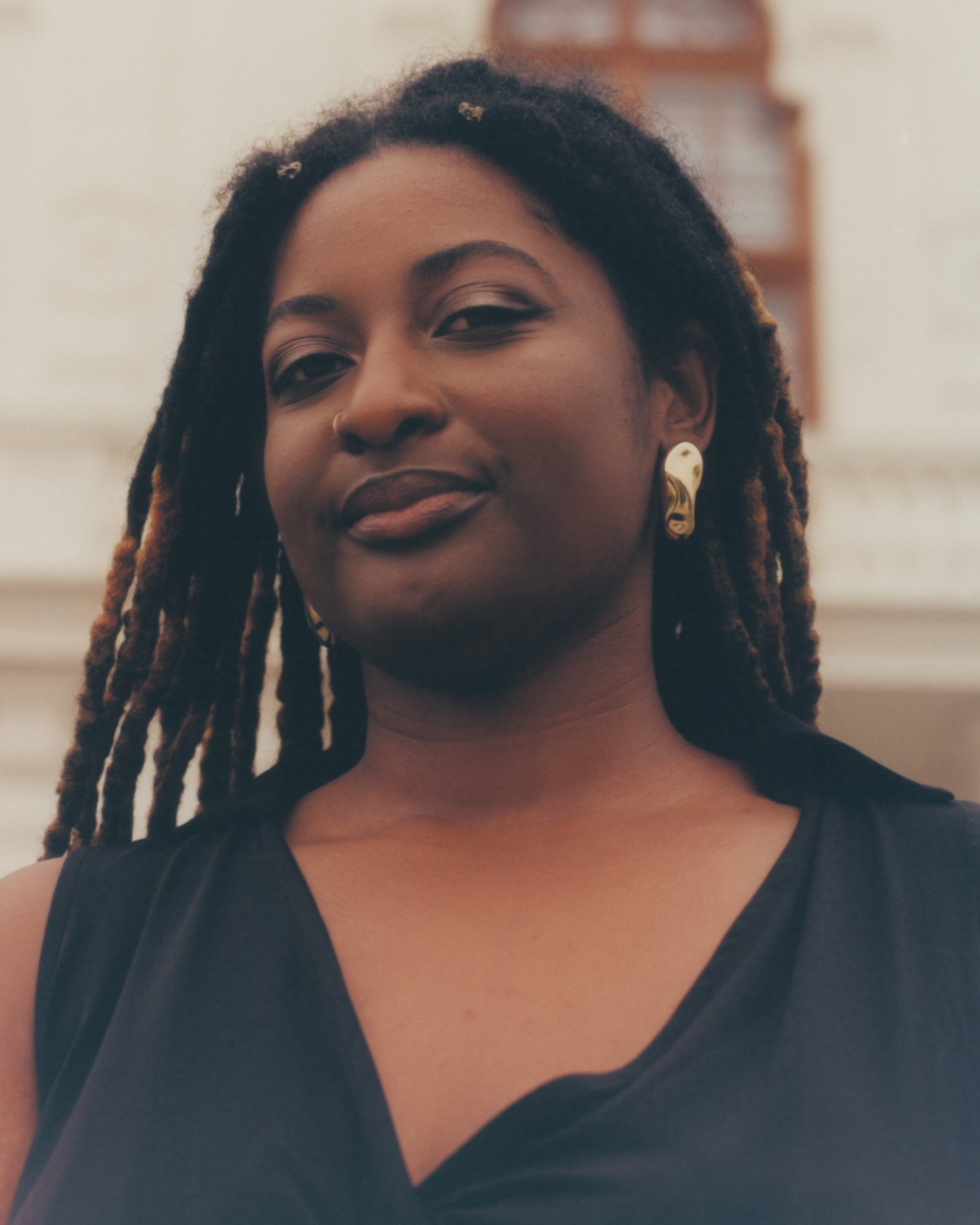 Tayla Myree with dark skin and long dreadlocks wearing a black sleeveless top and gold earrings, standing indoors in front of a blurred background.
