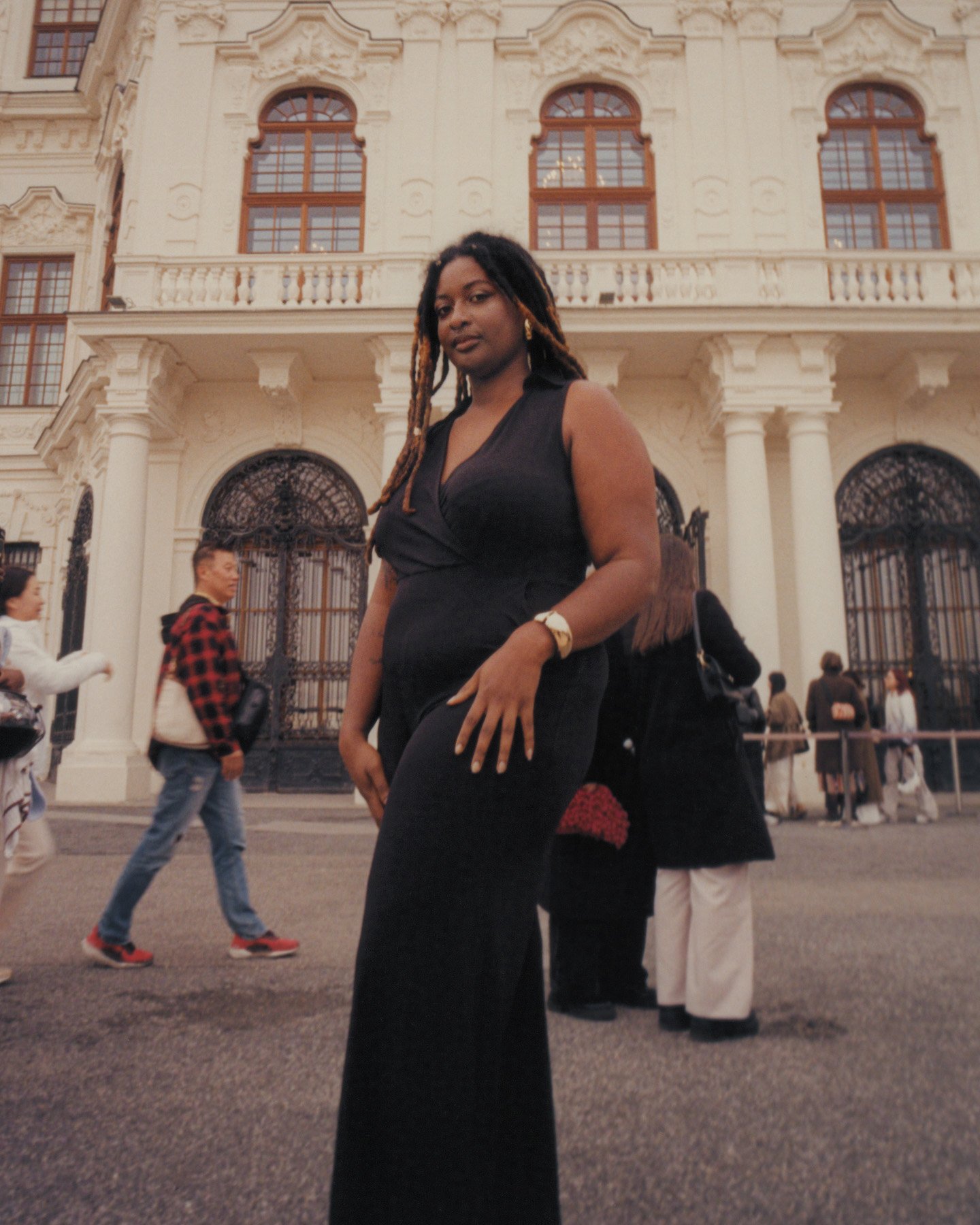 Tayla Myree with dreadlocks wearing a black sleeveless dress poses on a city street in front of an ornate historic building, with several pedestrians walking by in the background.