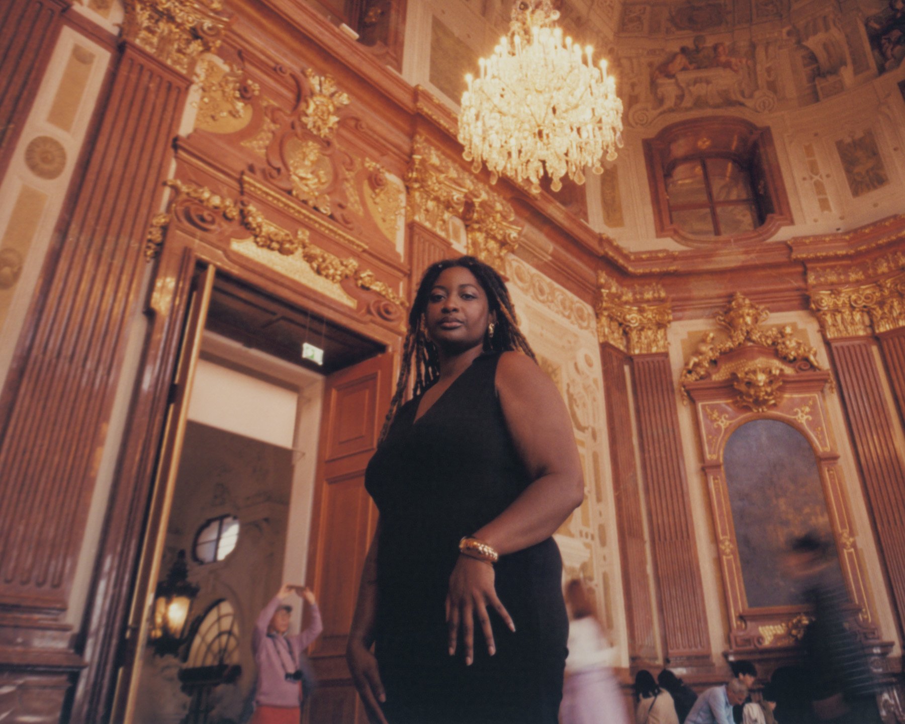 Tayla Myree with dreadlocks dressed in a black dress standing in a luxurious, ornate room with a large chandelier and intricate gold detailing on the walls