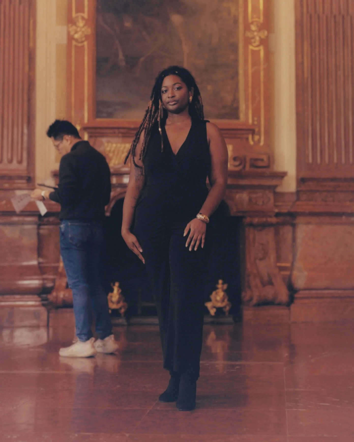 Tayla Myree in a black dress standing in front of a wooden wall in a grand hall, with a man in a black jacket and jeans in the background reading a paper.