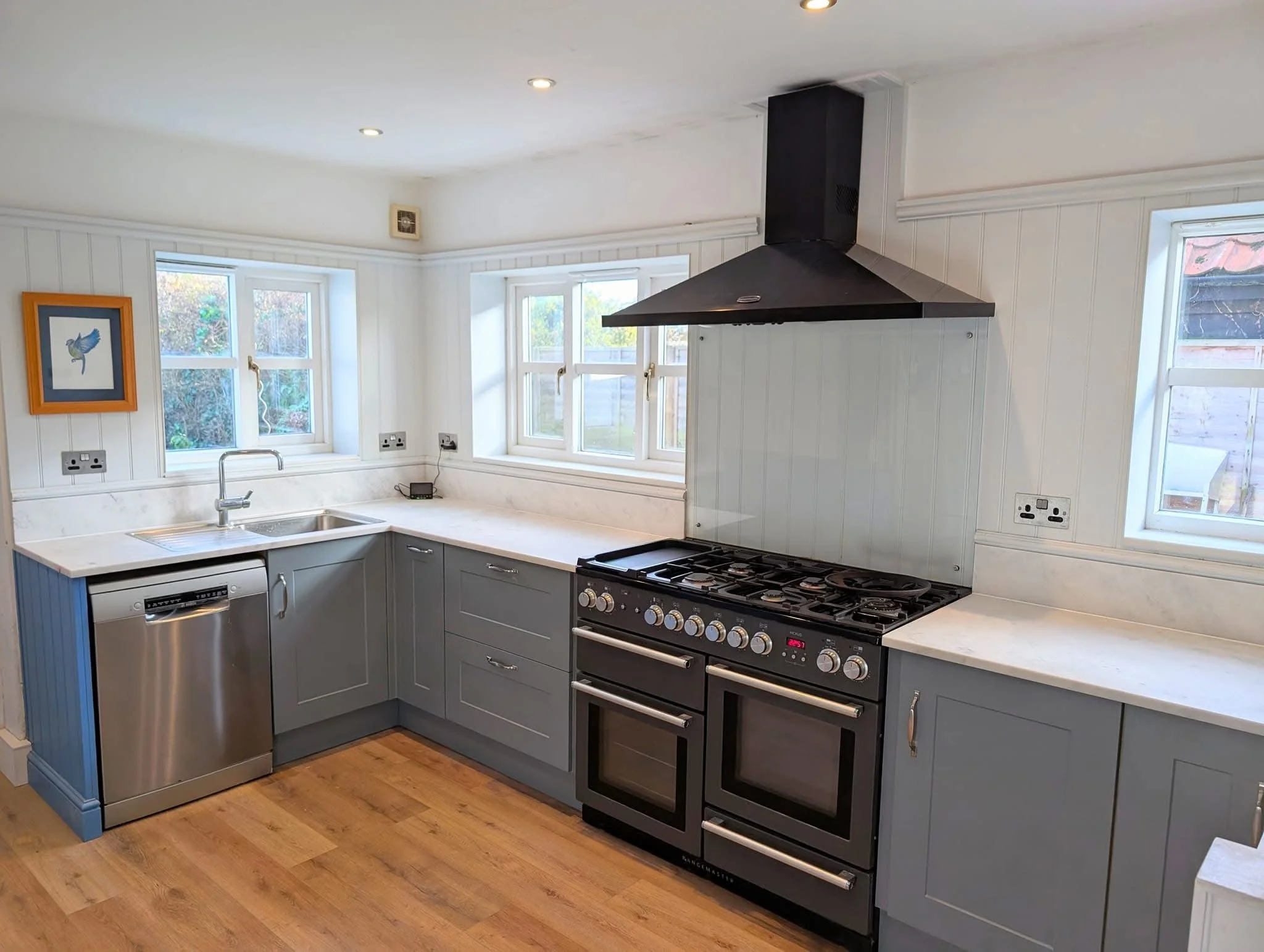 Kitchen with gray cabinets, gas stove, stainless steel dishwasher, white countertops, multiple windows, and a small framed artwork of a bird on the wall.