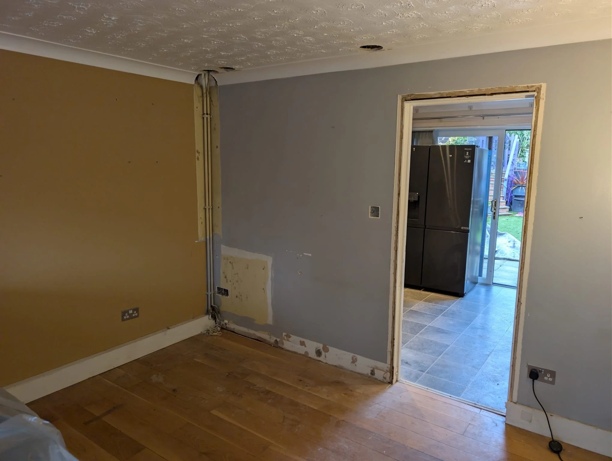 Interior room undergoing renovation with missing wall covering near doorway to kitchen, exposed pipes, and electrical outlets. The adjoining room has a black refrigerator and sliding glass door leading outside.