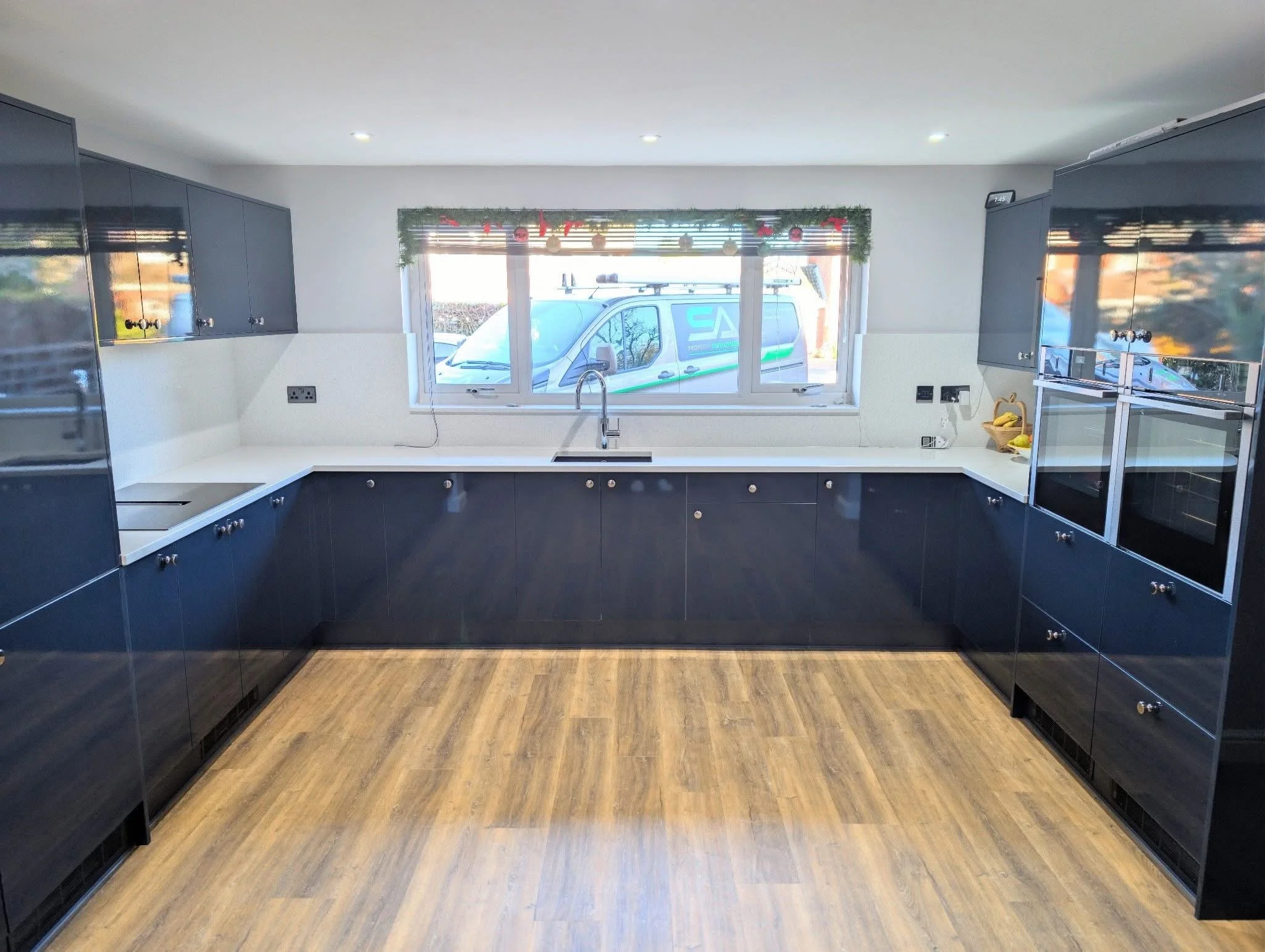Modern kitchen with dark lower cabinets, white countertop, and a large window above the sink with a view of a vehicle parked outside. The kitchen features built-in appliances, a wood floor, and some bananas on the counter.