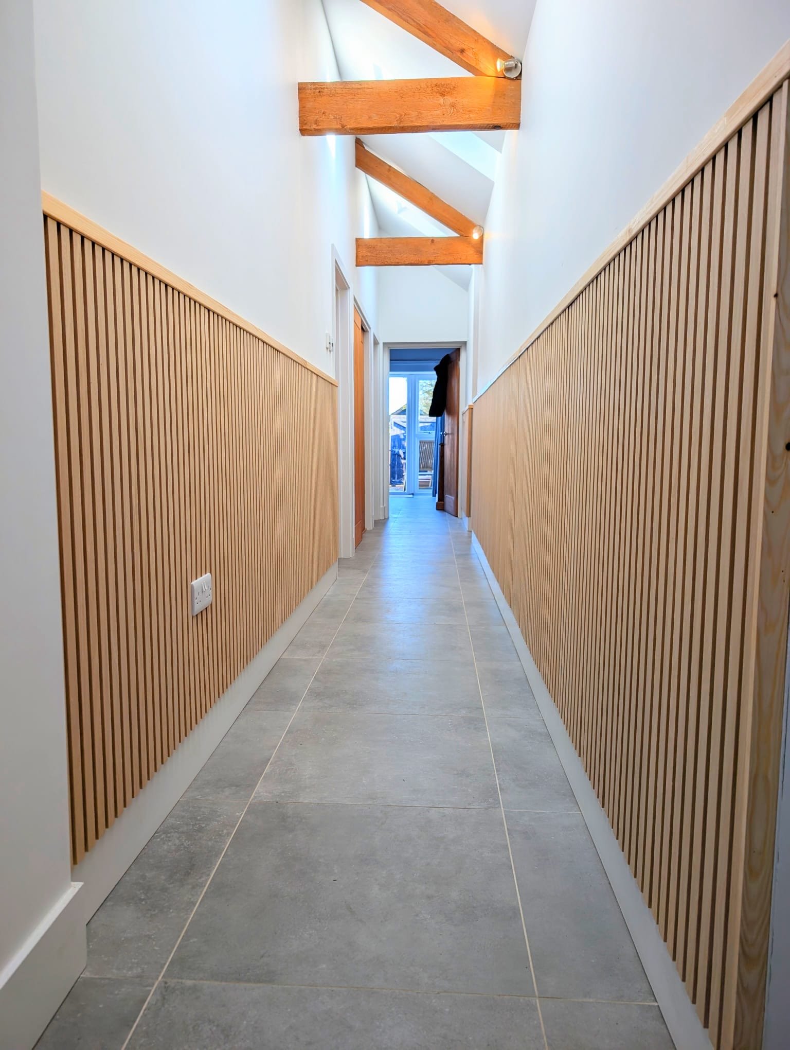 Empty hallway with wooden paneling on walls, ceiling with exposed wooden beams, and light-colored tiled floor, leading to a patio door.
