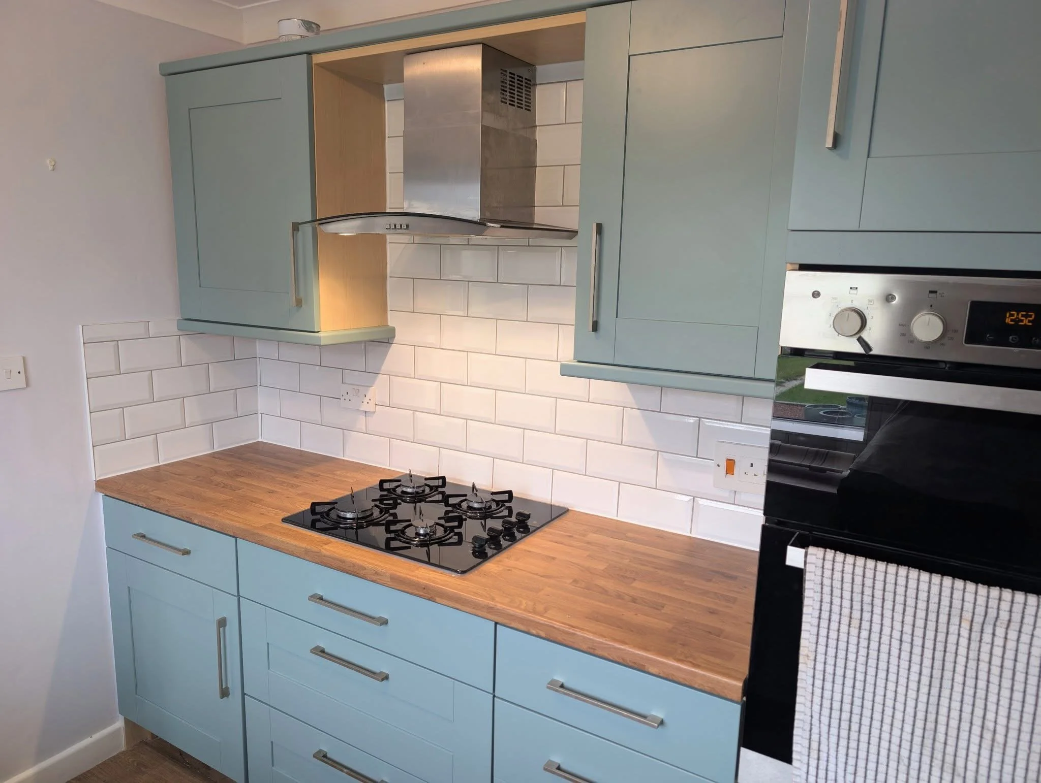 Kitchen with light blue cabinets, wood countertop, white subway tile backsplash, built-in gas stove, stainless steel range hood, and a black oven with a white tea towel hanging on its handle.